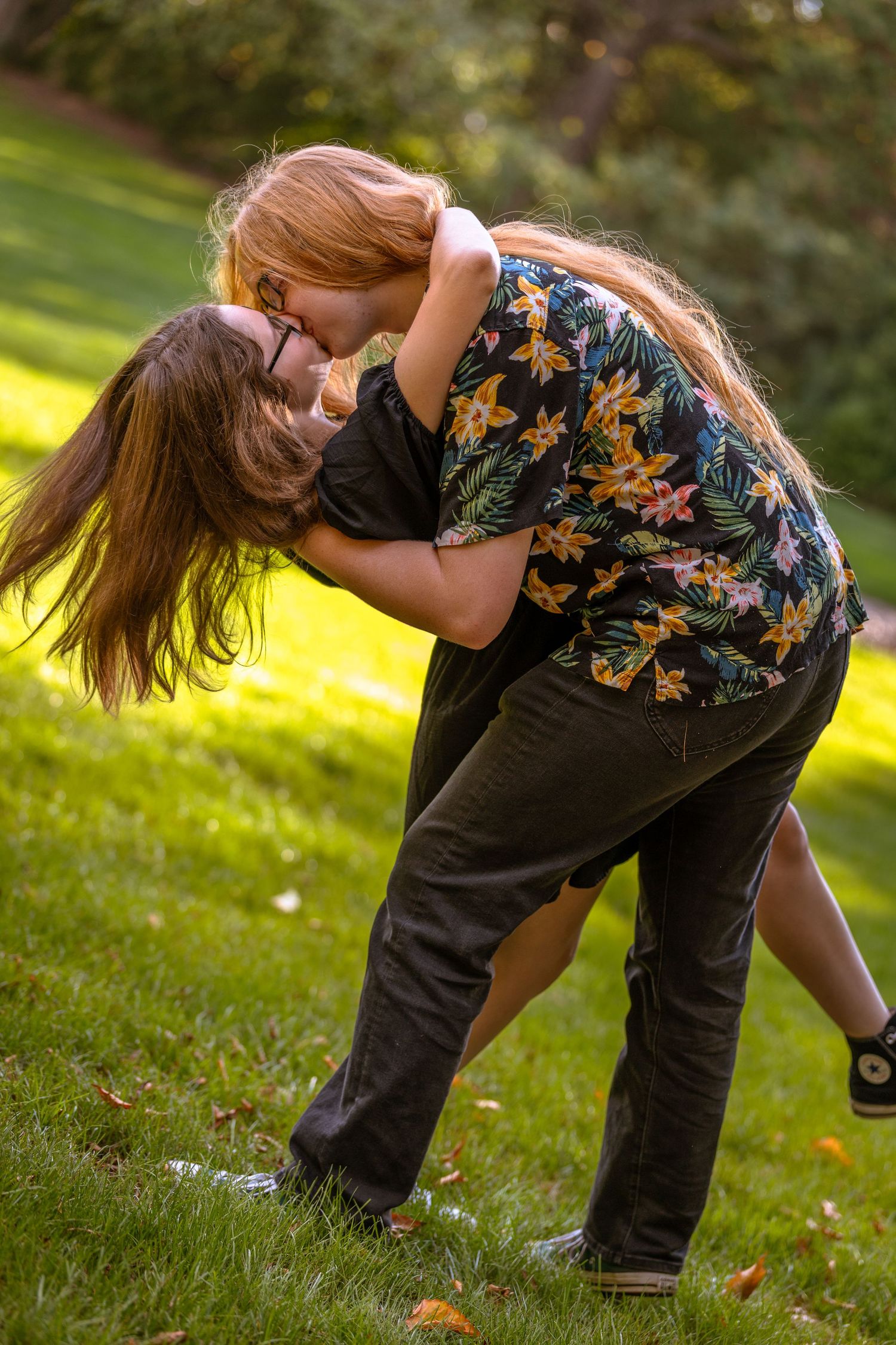 Two people playfully dip and dance together on green grass in a park setting.