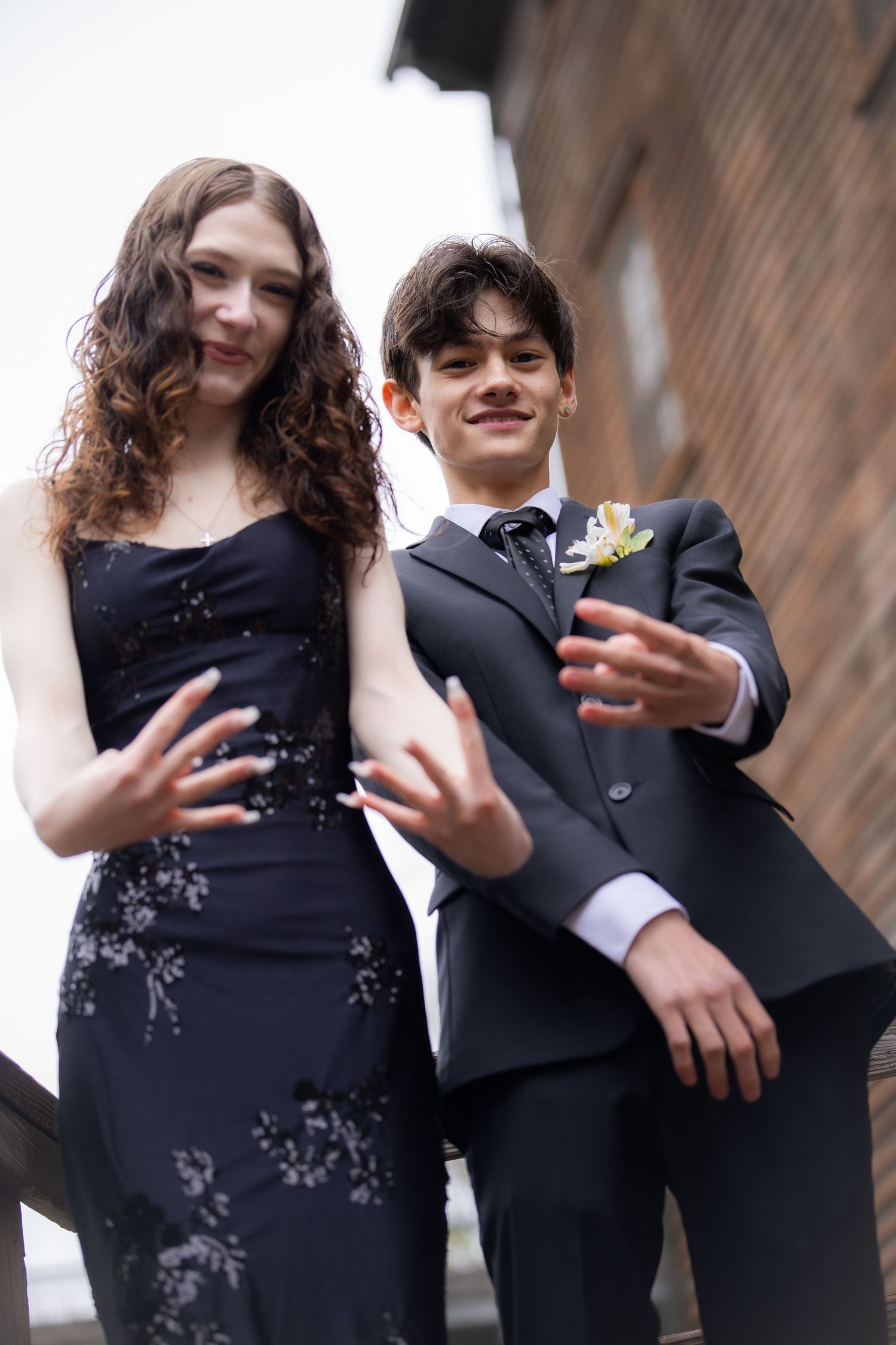 Two teens dressed in formal prom attire pose dramatically outdoors, wearing a black gown and gray suit with boutonniere.