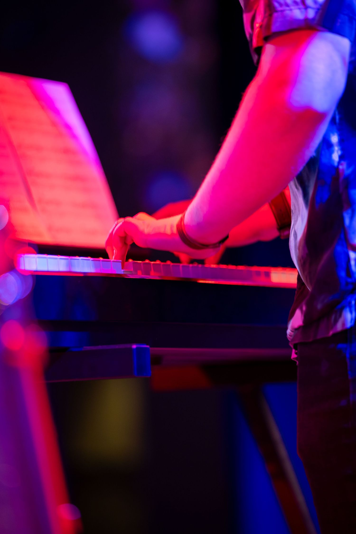 Musician playing keyboard synthesizer on stage under vibrant red and purple concert lighting during live performance.