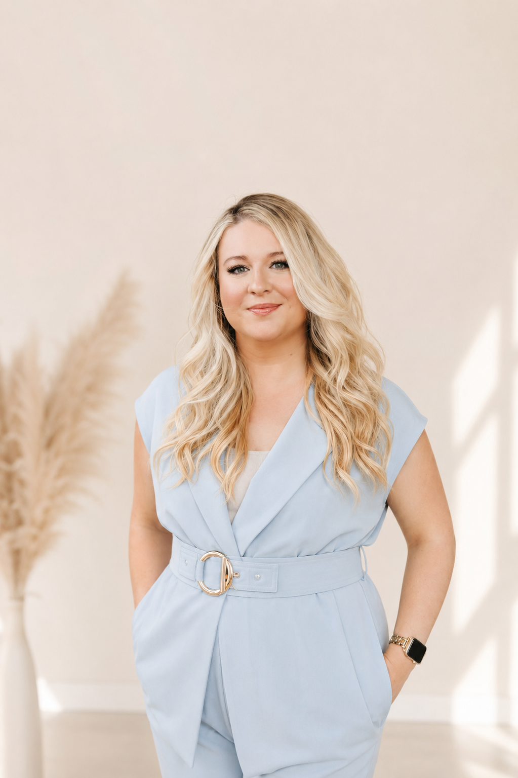 Smiling blonde woman in a light blue belted jumpsuit posing in a bright studio setting.