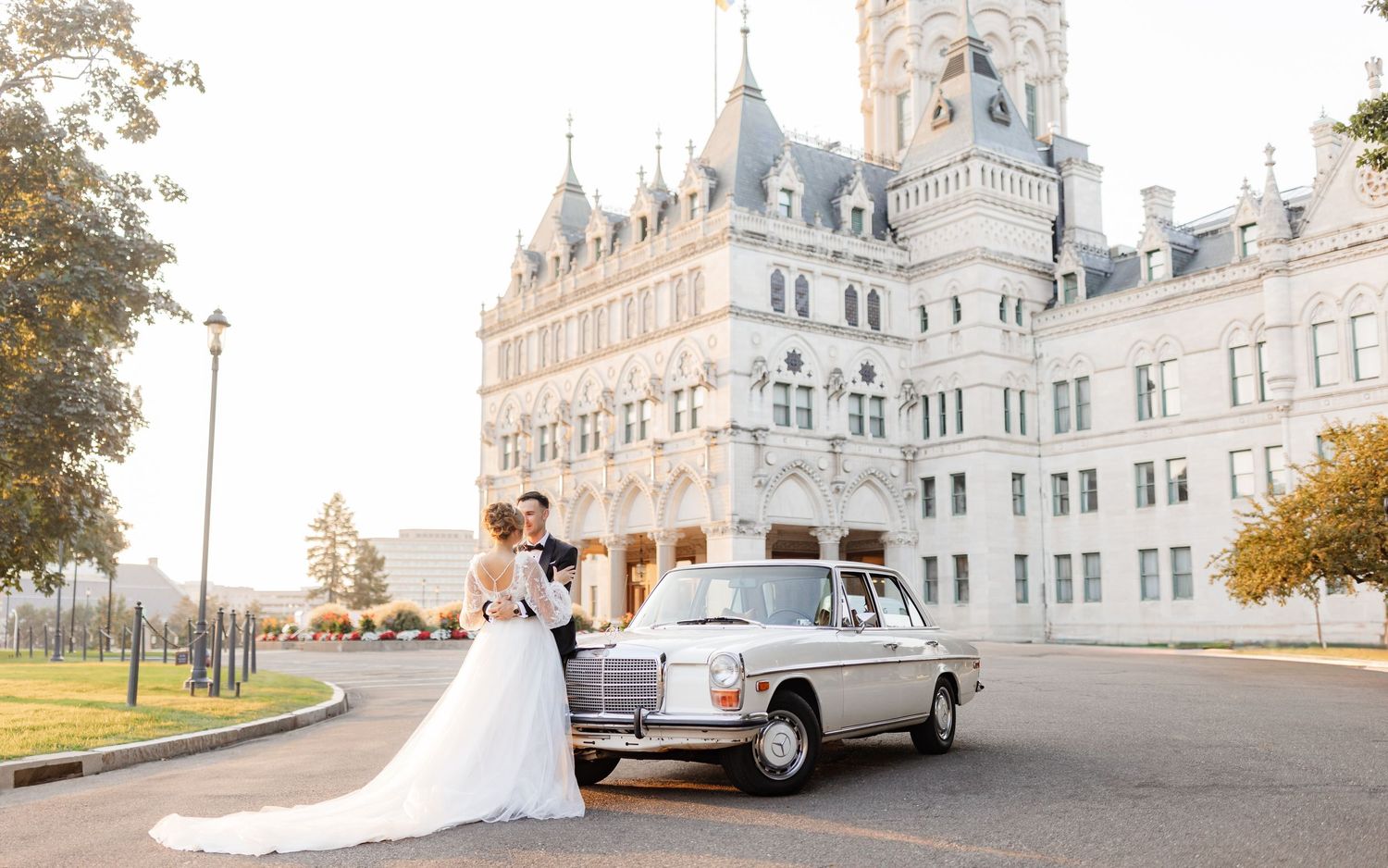 Bride and groom pose beside a vintage Mercedes-Benz in front of an elegant château-style building on their wedding day.