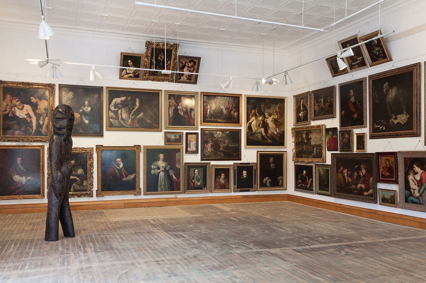 Classical paintings displayed salon-style on gallery walls with visitor viewing artwork.