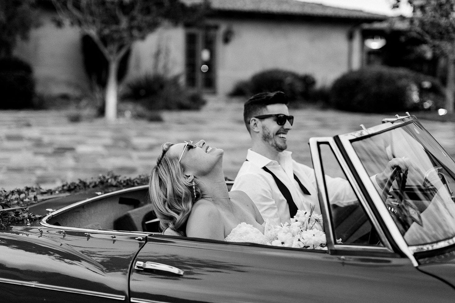 A couple laughs together while riding in a classic convertible car on a sunny day.