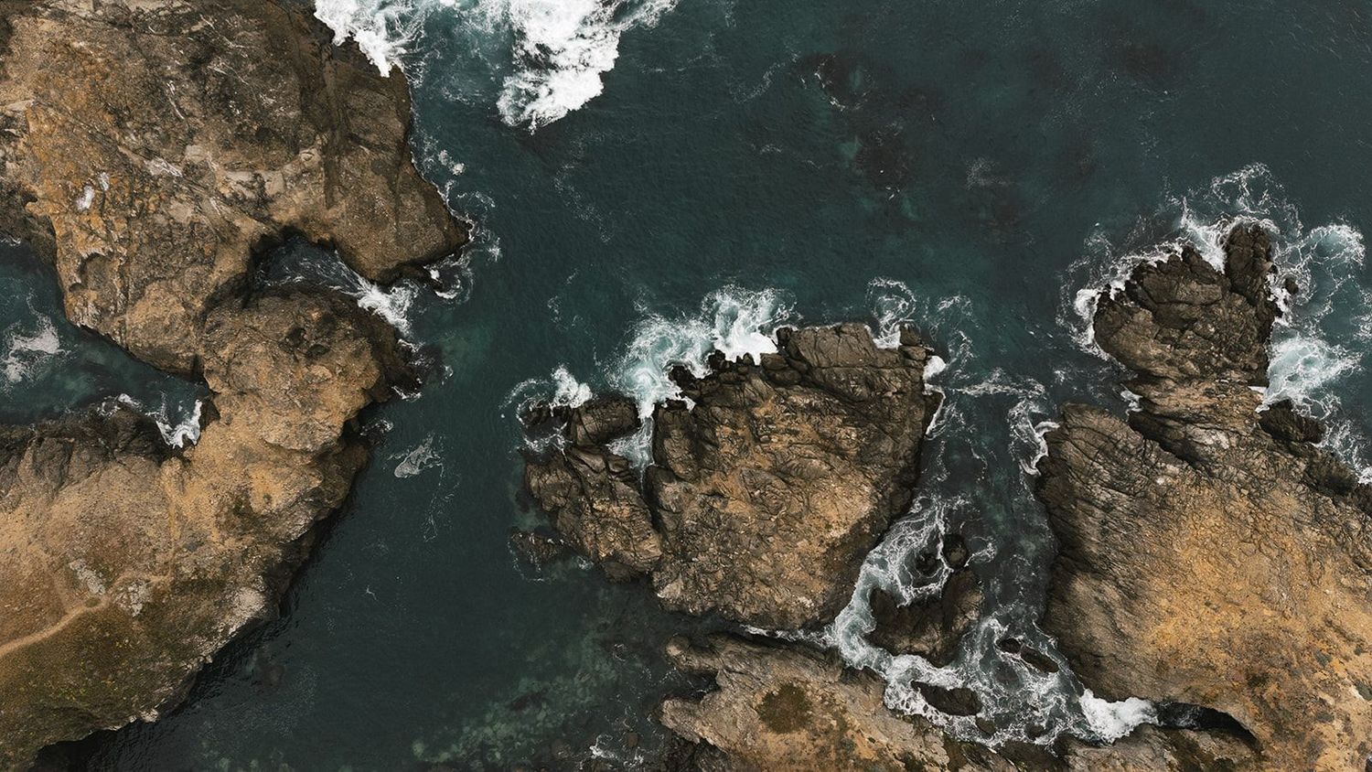 Aerial view of a coastline with rocky outcrops and white surf against turquoise waters.