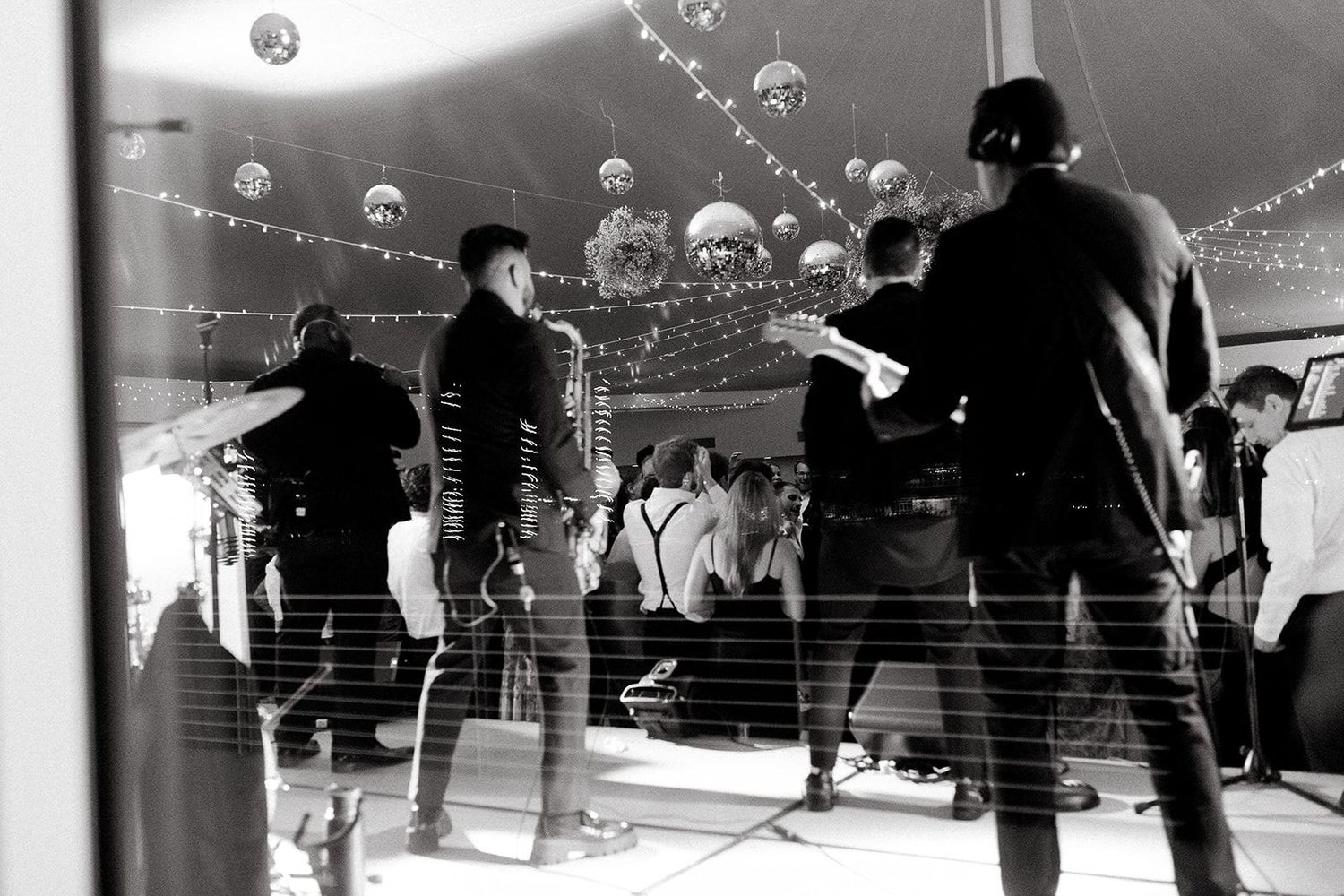 Black and white photo of a band performing on stage at a wedding reception under string lights.