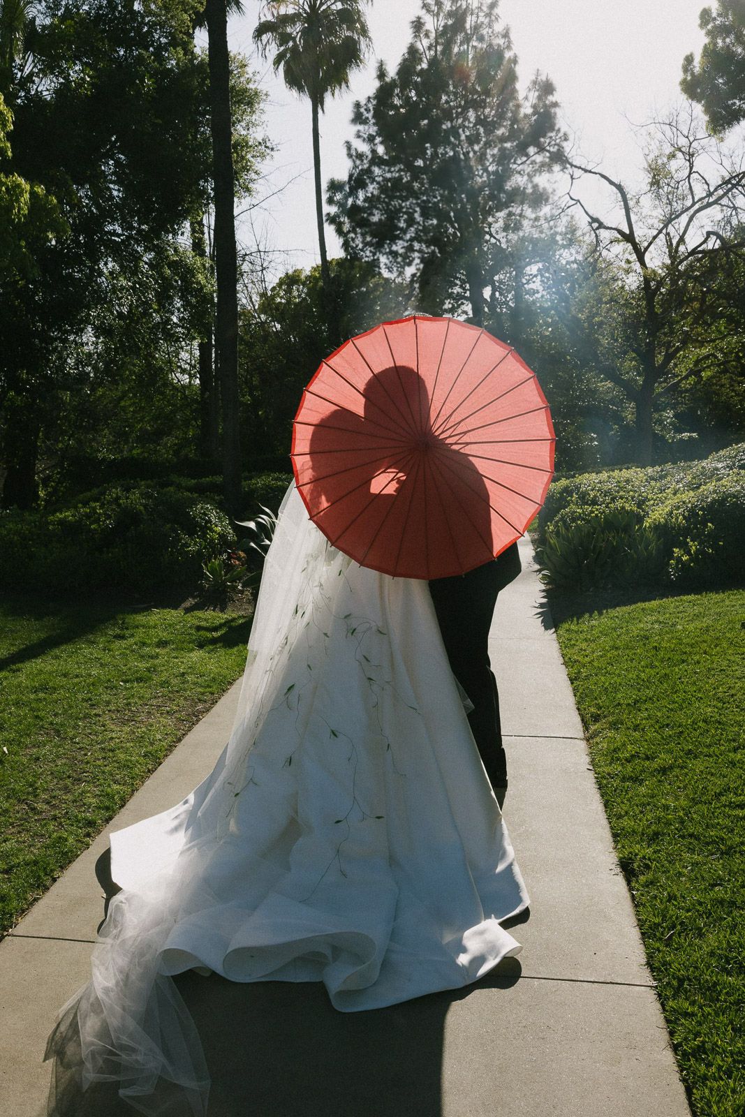 Wedding dress and coral parasol photographed along a garden pathway.