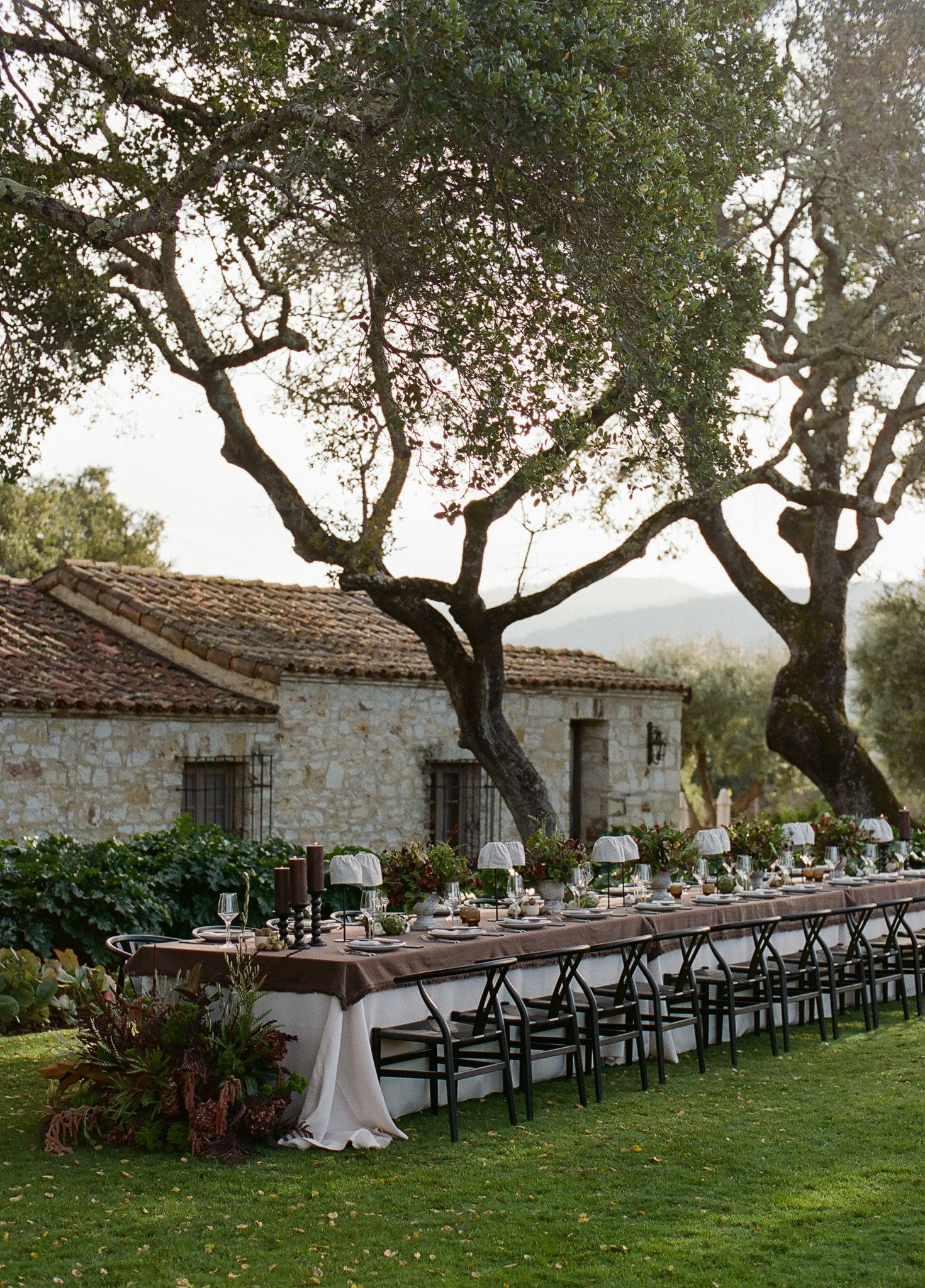 Wedding reception setup with long tables and black chairs on lawn by stone building.