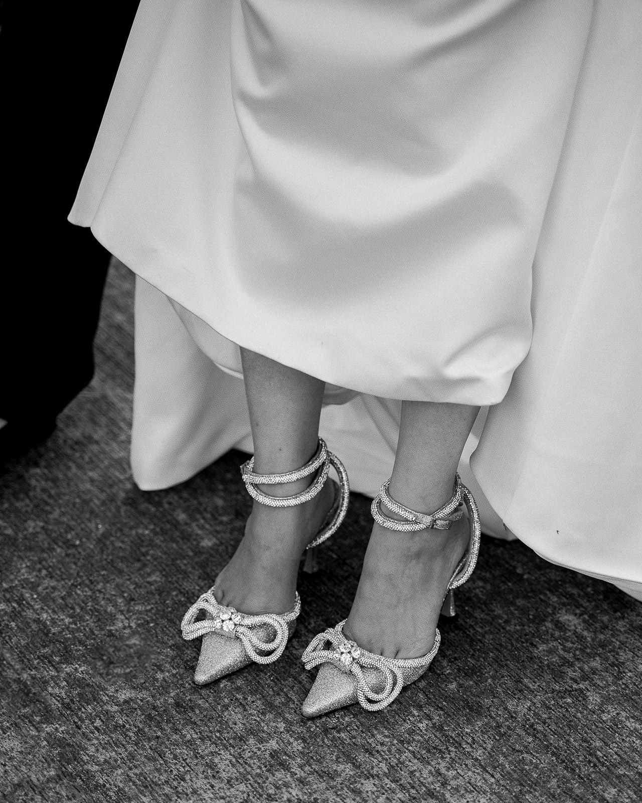 Close-up of white bridal shoes with delicate bow details on a textured surface.