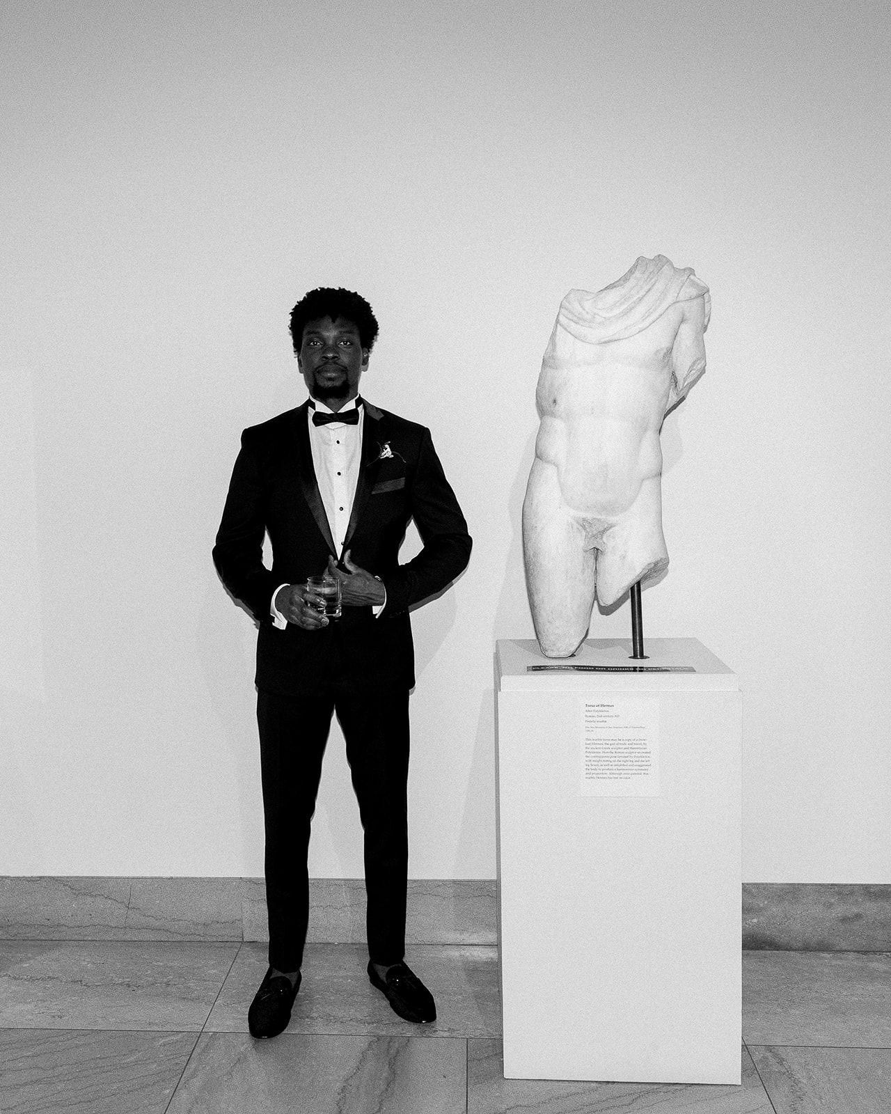 A formal portrait of wedding guest at Legion of Honor in a tuxedo standing next to a classical statue on display.