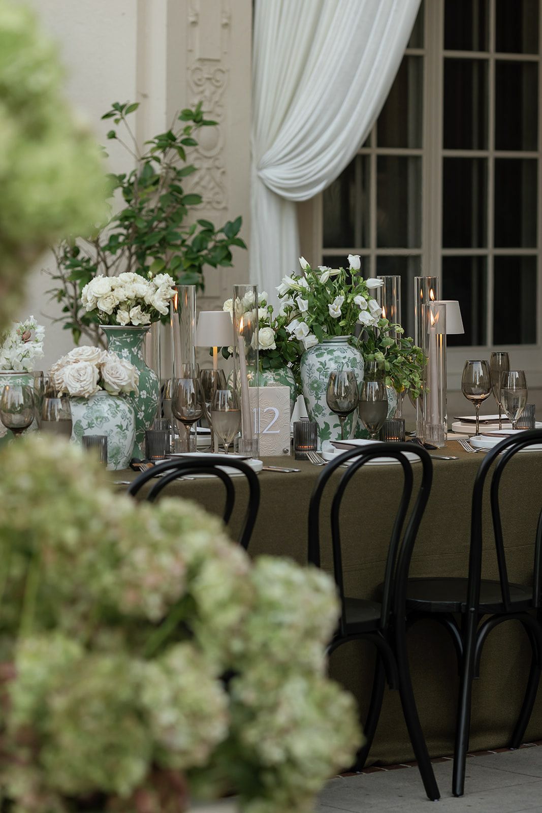 Elegant wedding reception tables with white floral centerpieces and black chairs in a light-filled venue.