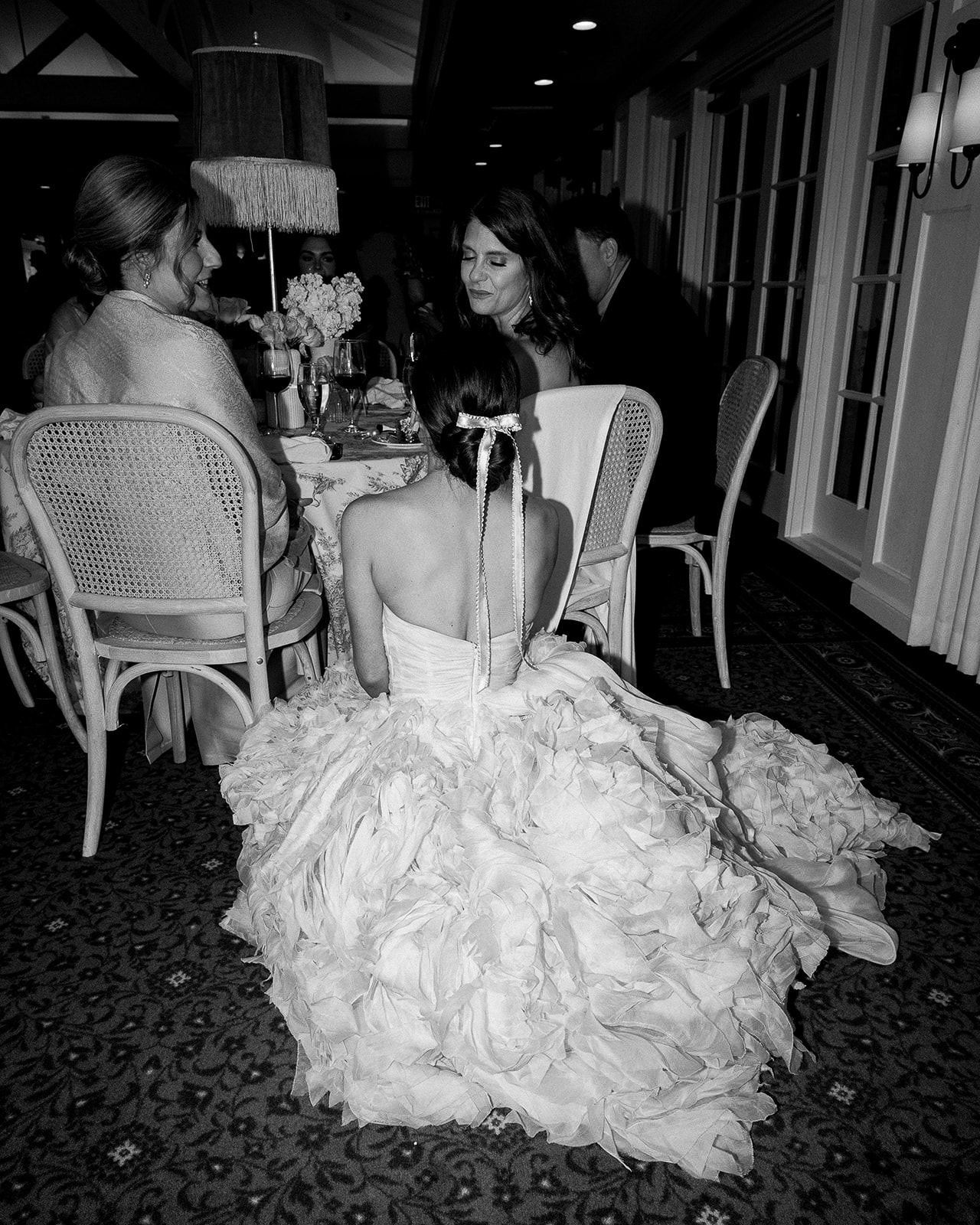 Black and white photo of a ruffled wedding dress cascading over a dining chair at a reception.