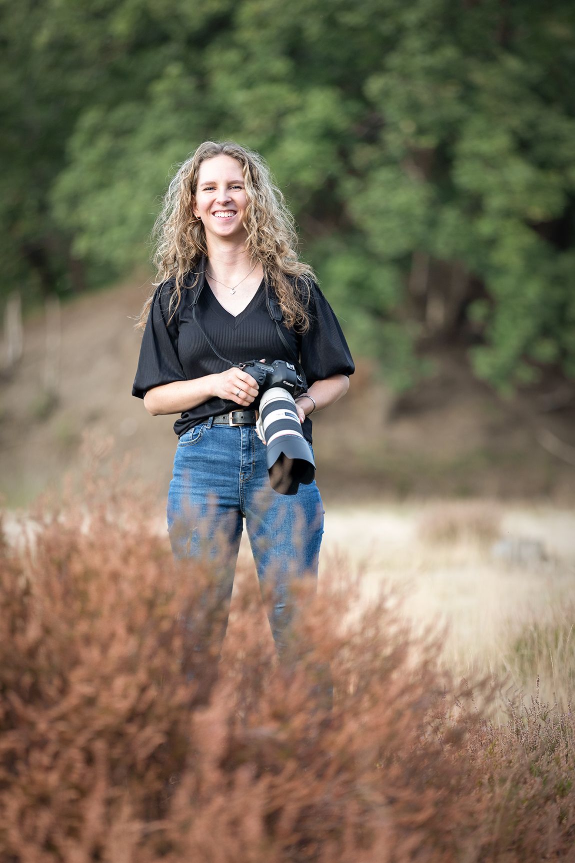 A photographer poses with camera equipment in a field of dried grass during an outdoor photo session.