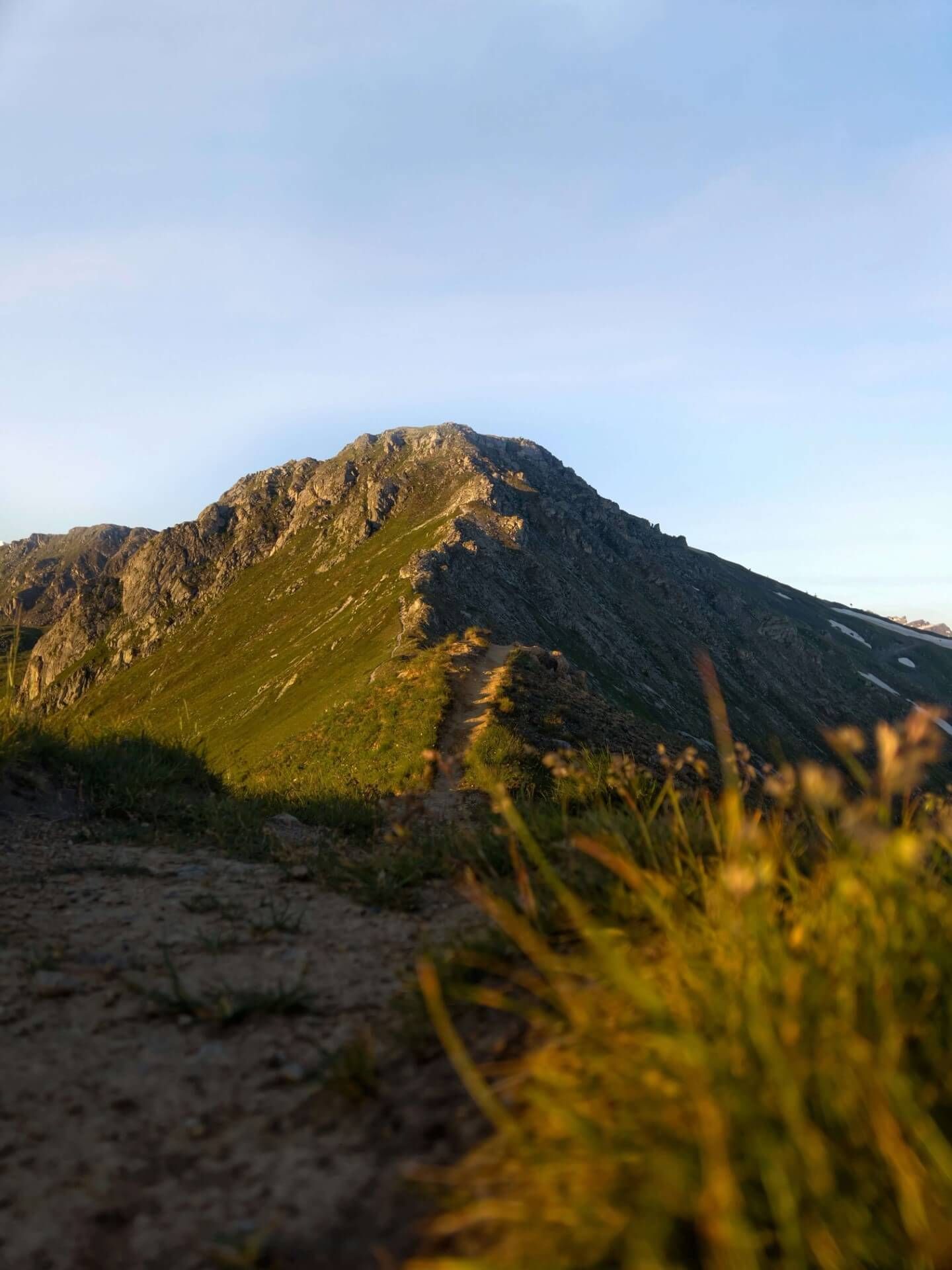 Von goldenem Sonnenlicht beleuchtete Felsgipfel mit einem Wanderweg und Gras im Vordergrund.
