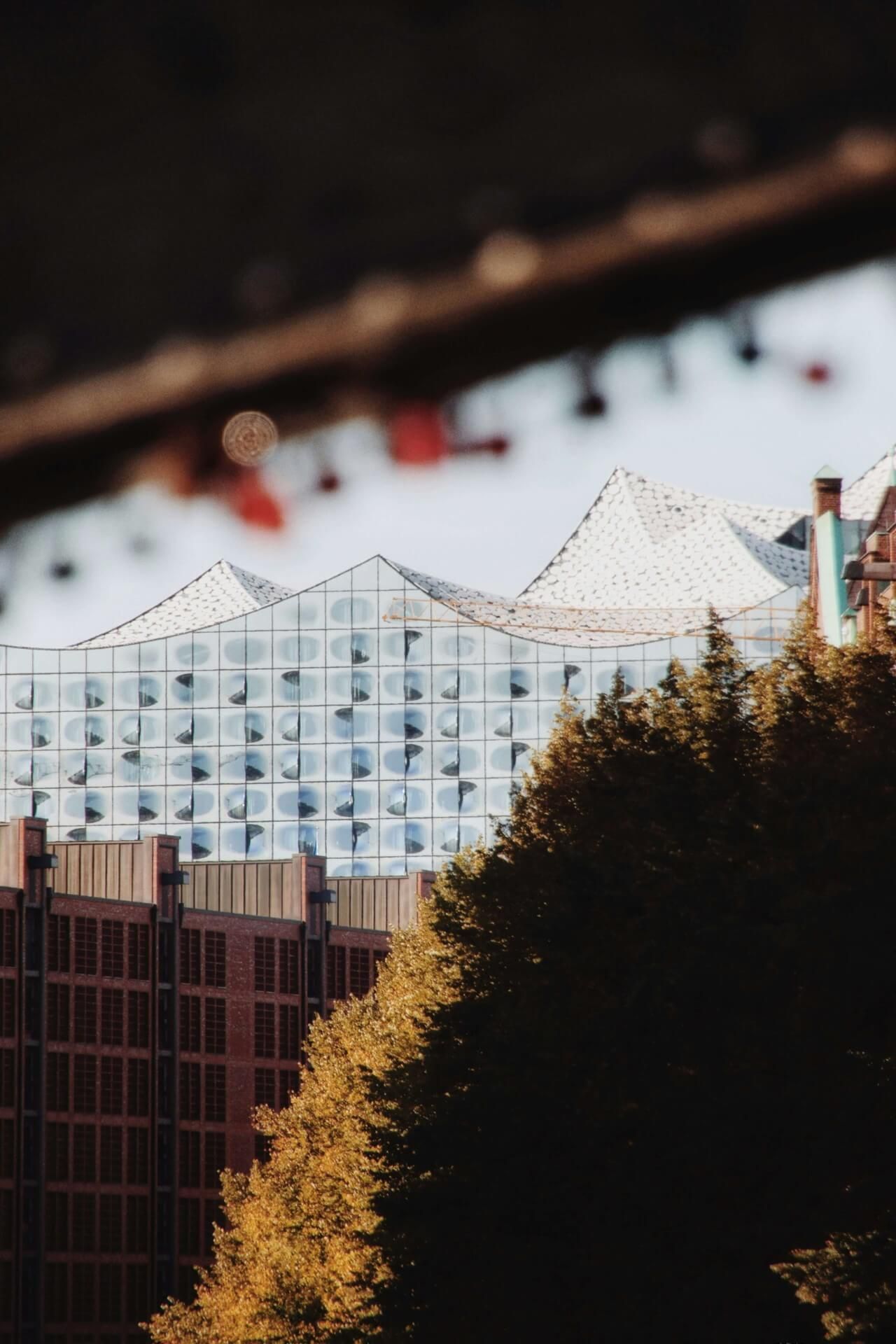 Moderne Glasfassade der Hamburger Elbphilharmonie mit Herbstbäumen, roten Backsteinhäusern und einem Brückenpfeiler im Vordergrund.