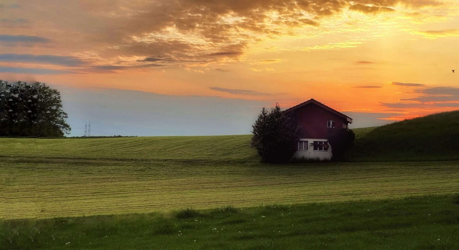 Rot-braune Scheune vor einem dramatischen orangefarbenen Sonnenuntergangshimmel über ländlichem Ackerland.