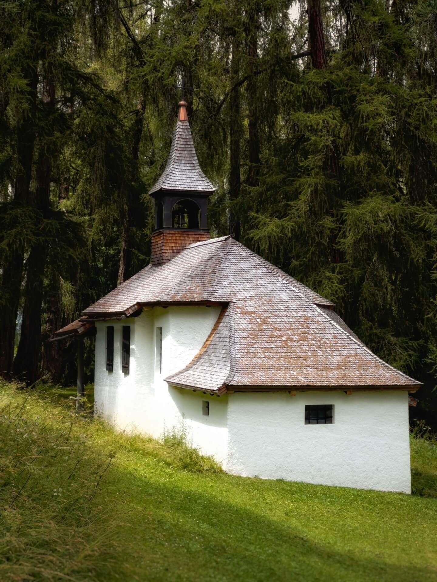 Kleine weisse Kapelle mit spitzem Turm inmitten eines dichten immergrünen Waldes.