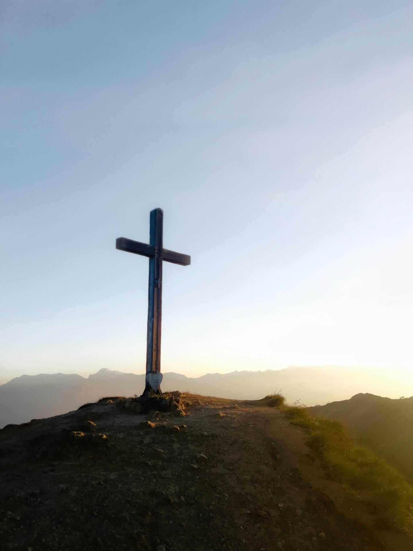 Silhouette eines grossen Gipfelkreuzes auf einer Bergkuppe vor einem leuchtenden Sonnenaufgangshimmel.