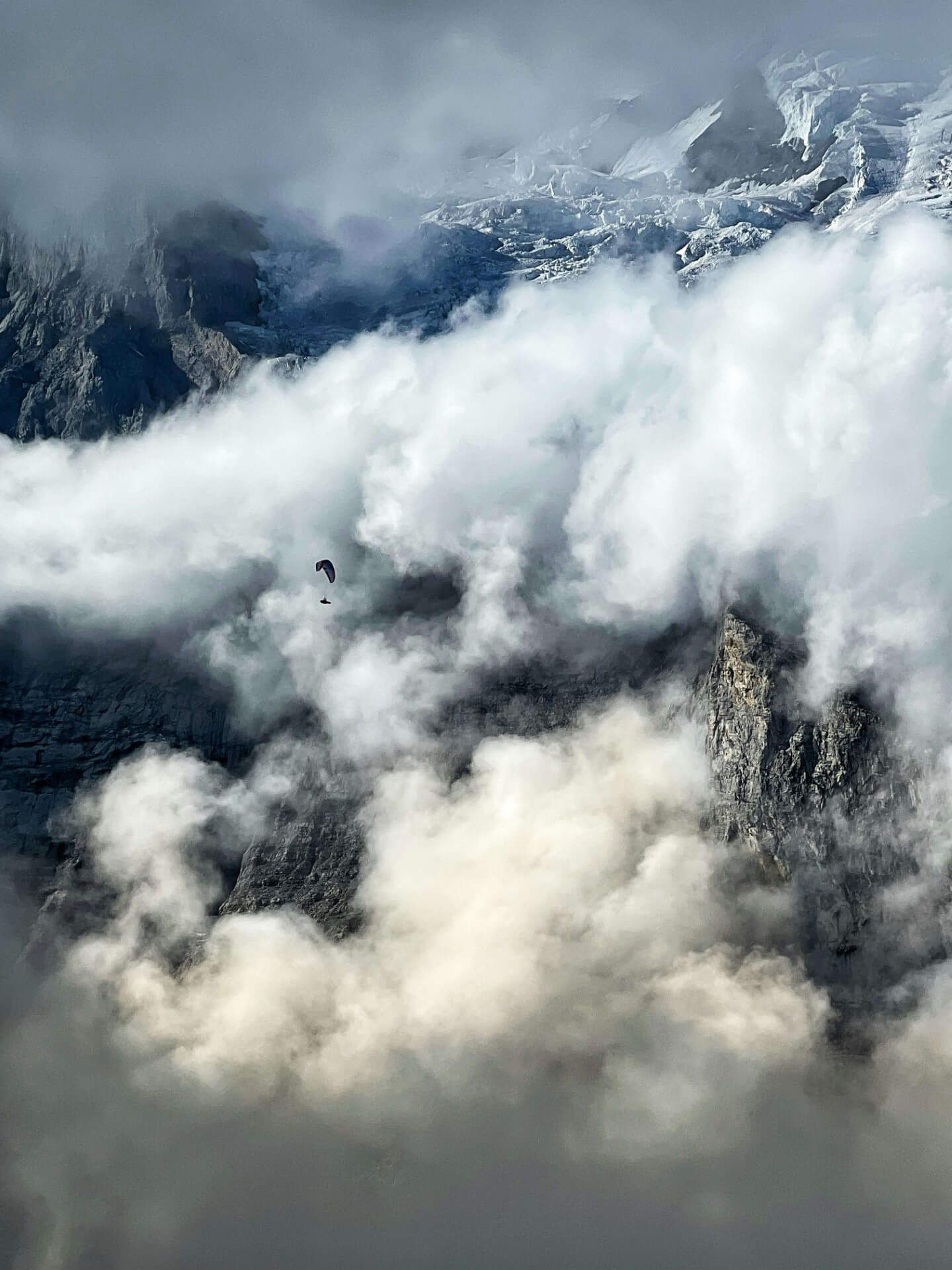 Ein Gleitschirm, klein im Bild, fliegt durch riesige, dramatische Wolkenformationen vor einer Felskante mit Gletscher im Hintergrund.