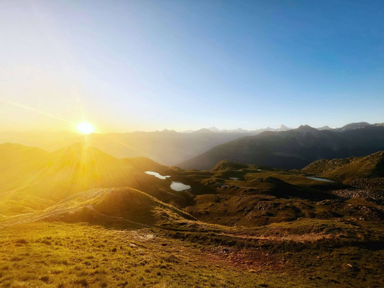 Panoramablick auf die Berge bei Sonnenaufgang mit goldenem Licht, das über die sanften Hügel fällt, mit wolkenlosem Himmel im Hintergrund.