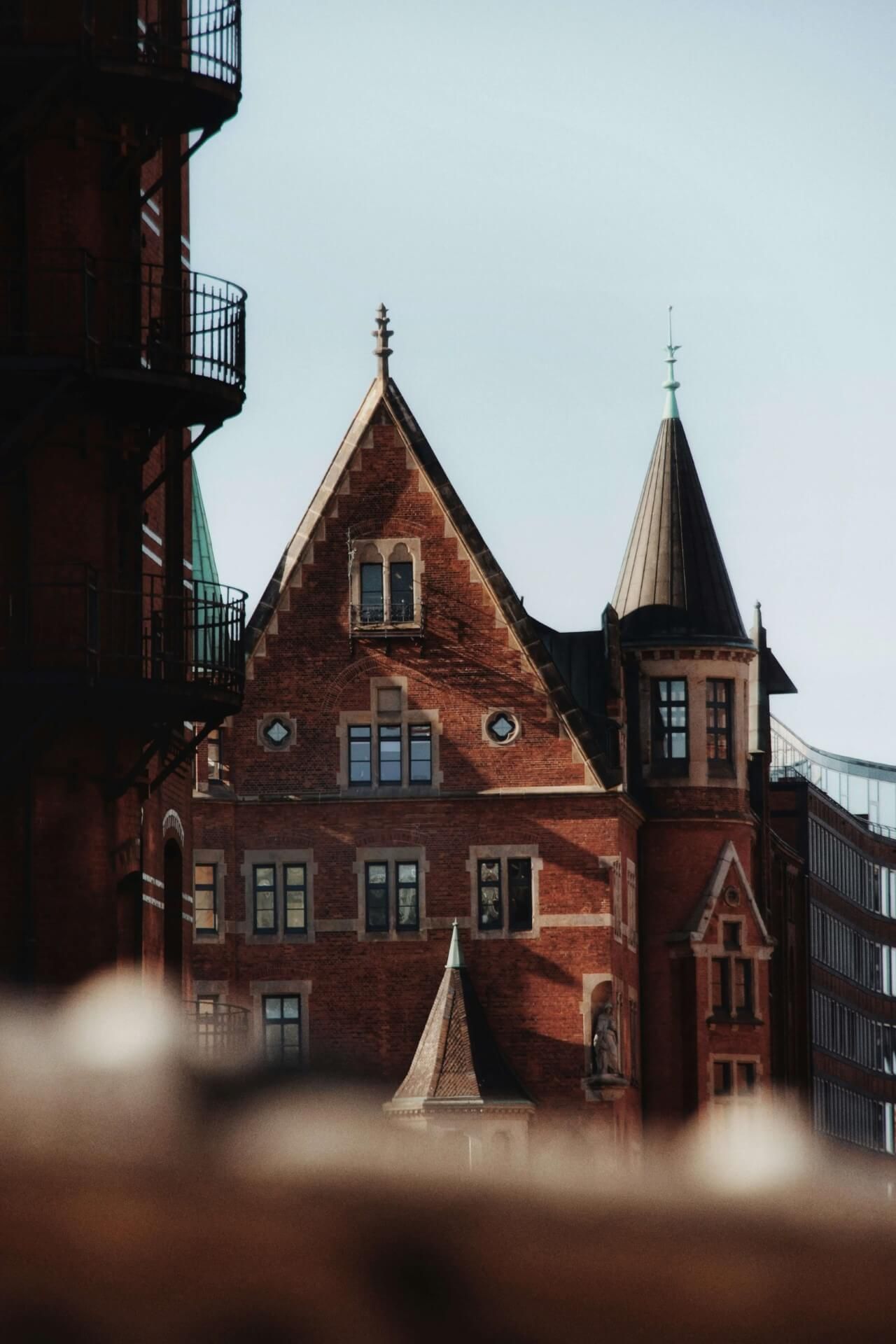 Rote Backsteinarchitektur im gotischen Stil mit verschnörkelten Turmspitzen und Türmchen vor einem blassen Himmel in Hamburg.