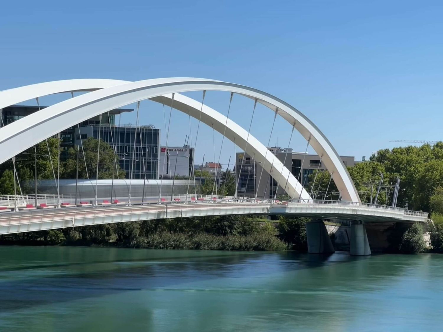 Moderne weisse Bogenbrücke überspannt türkisfarbenes Flusswasser an einem sonnigen Tag, während ein Tram darüberfährt.