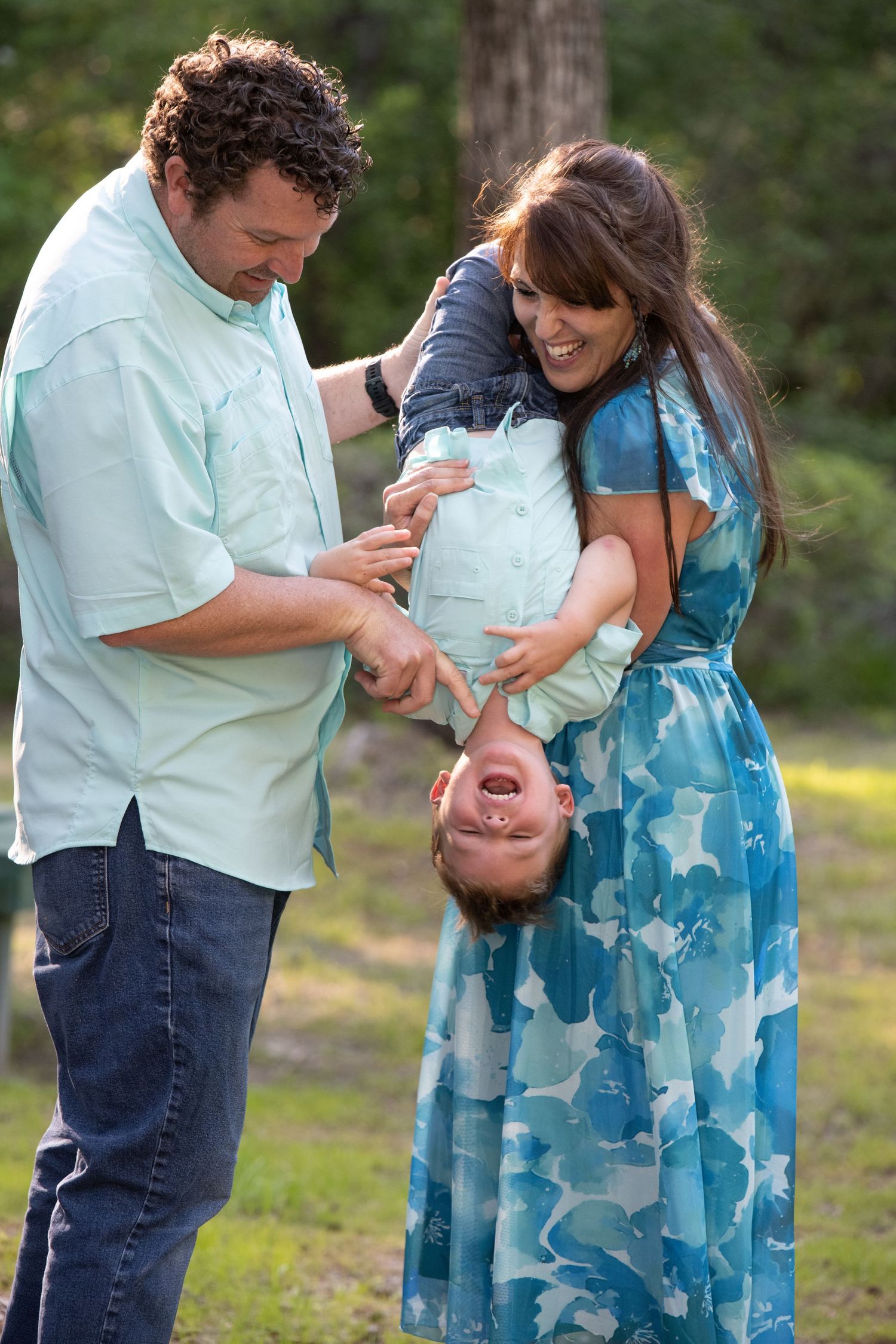 Happy family playing outdoors, parents holding toddler upside down, laughing together in a green park setting.