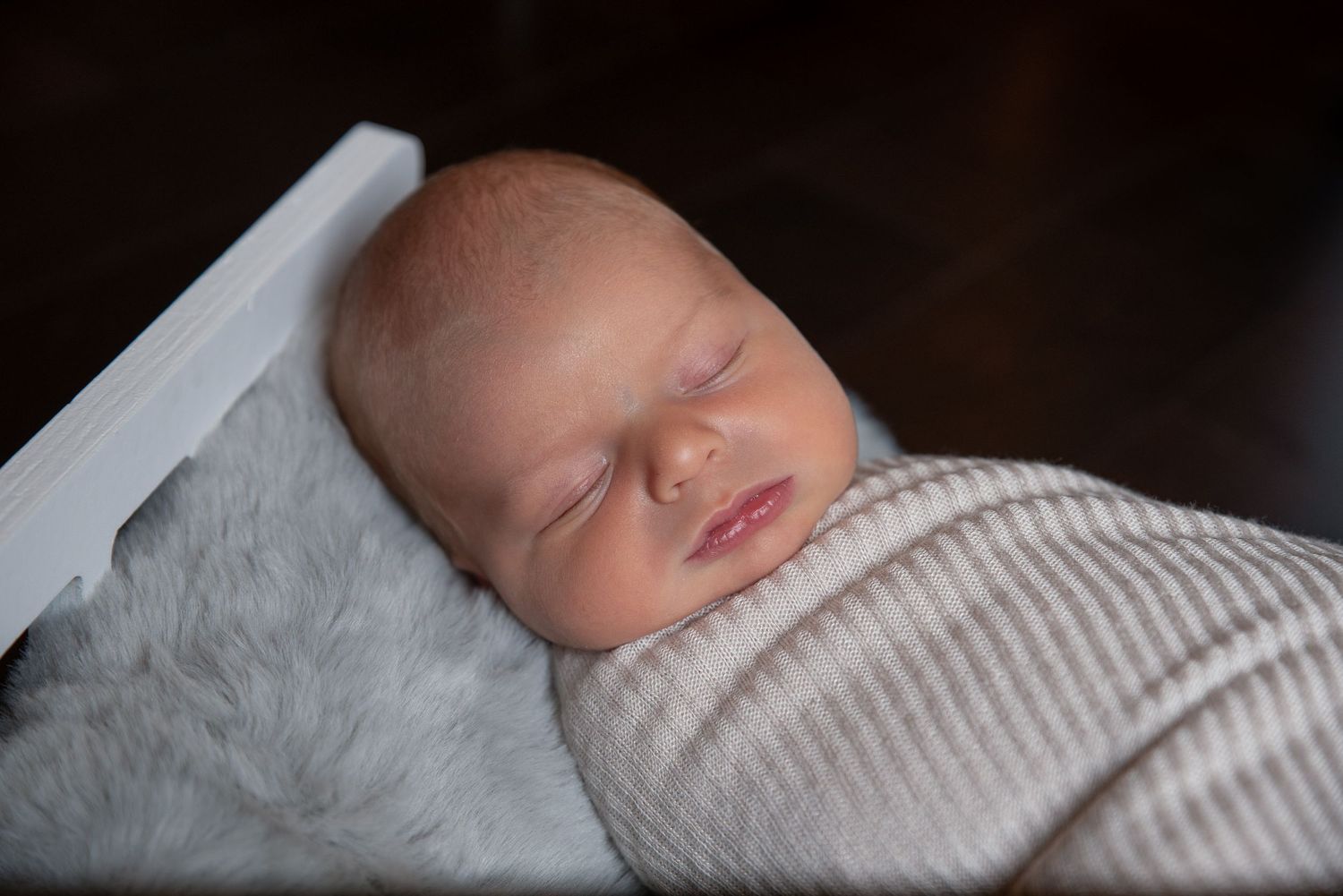 Peaceful newborn baby swaddled in a cream knit wrap, sleeping on a soft gray fur blanket in a white wooden bed.