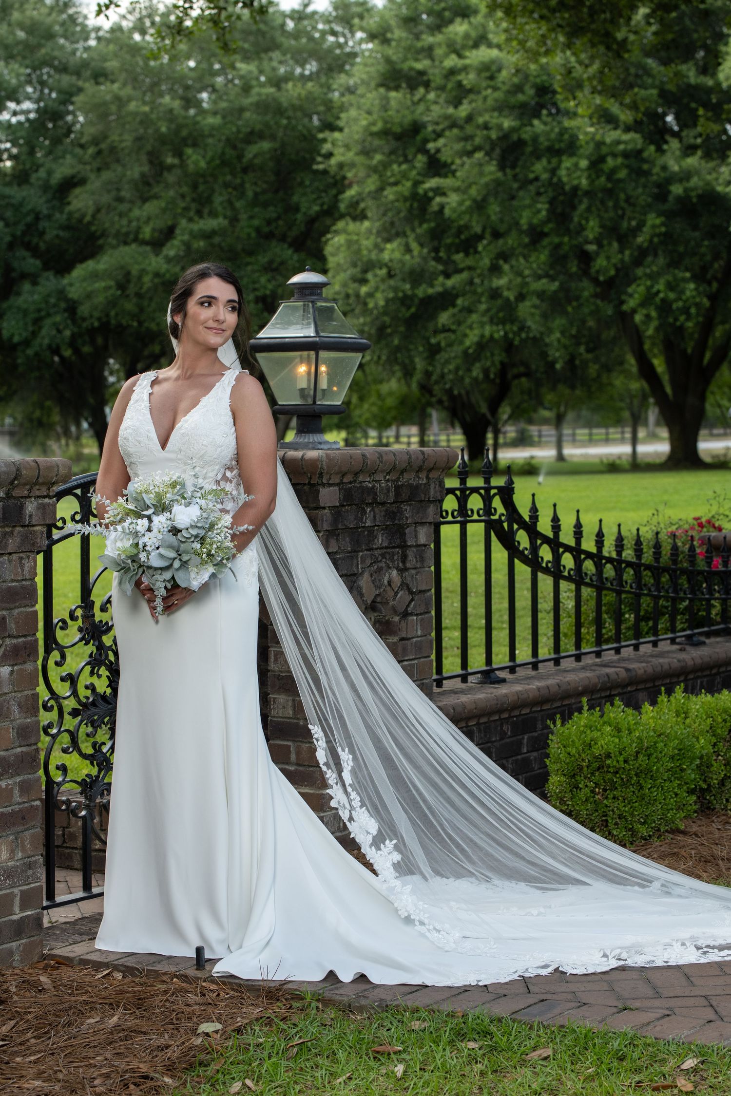 Bride in white gown with lace veil holding bouquet, standing by iron fence and stone wall in lush garden setting.