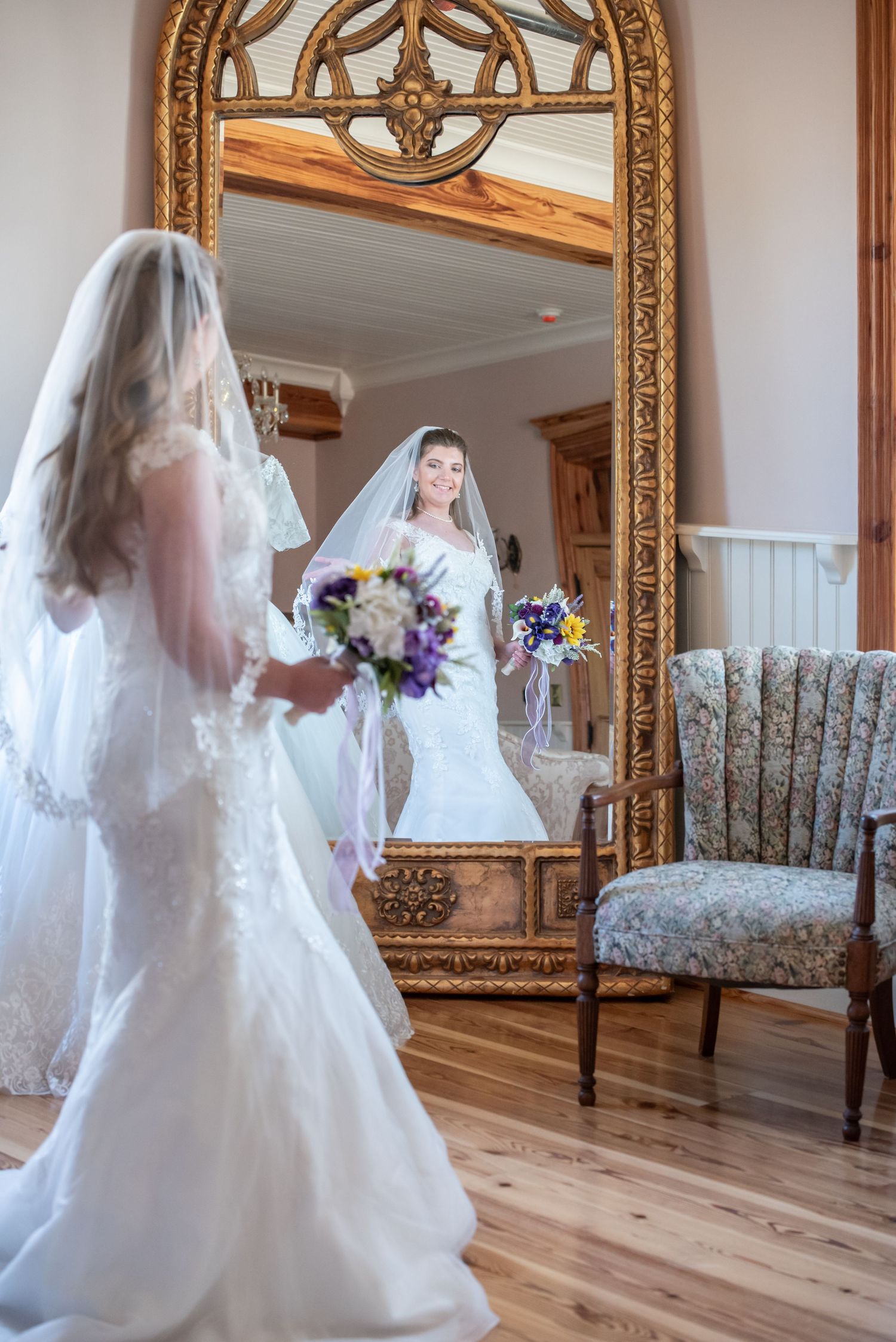 A bride in a white wedding gown with veil stands before an ornate gold-framed mirror holding a purple floral bouquet.