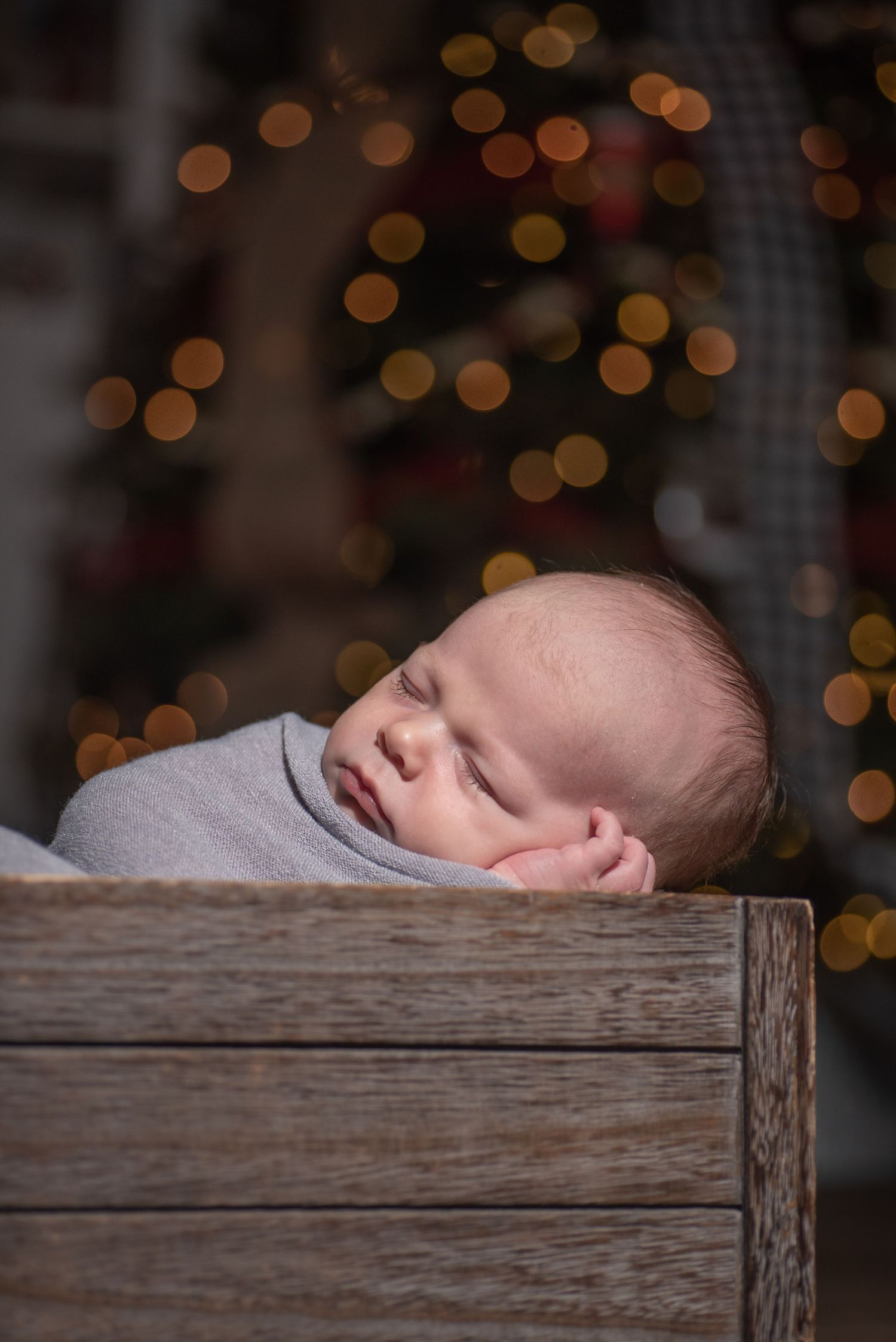 Sleeping newborn baby in wooden crate with soft bokeh Christmas lights in background.