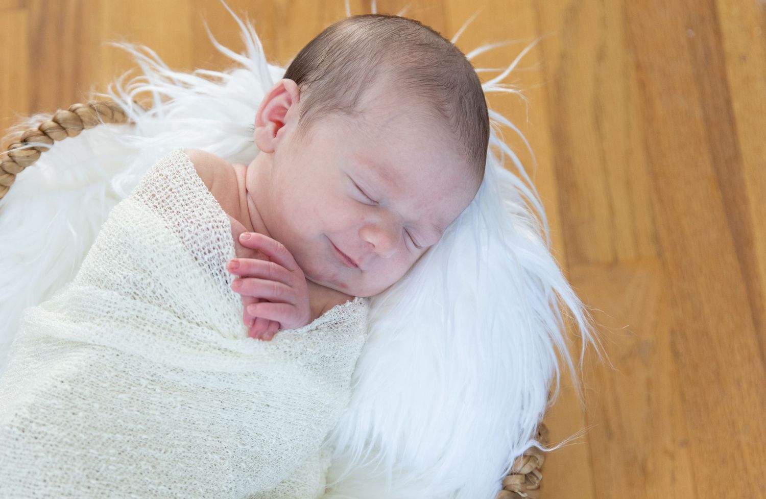 A peaceful sleeping newborn wrapped in a white blanket rests on a soft white fur blanket against a wooden floor.