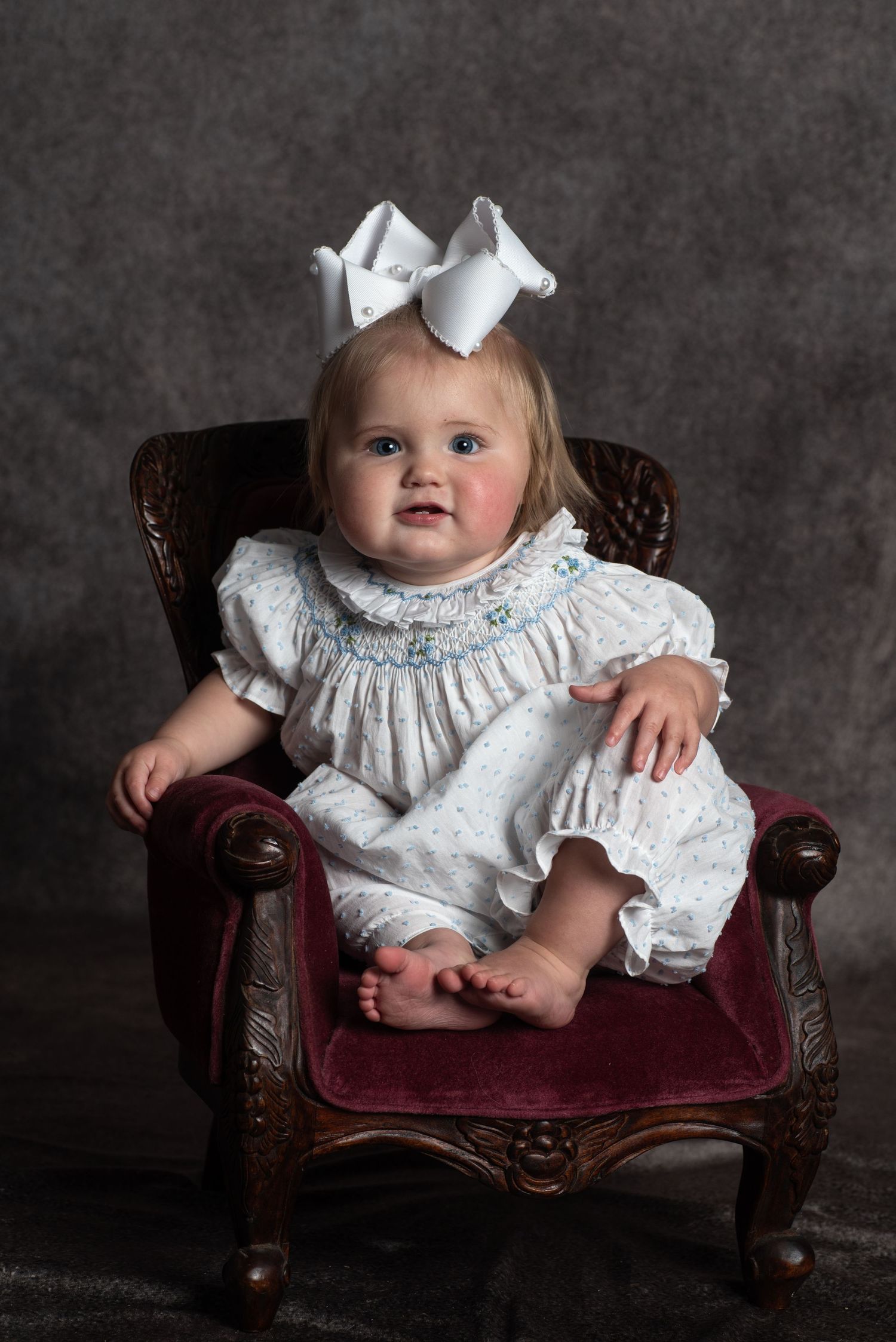 A little one in a white smocked dress and bow sits in an ornate vintage burgundy leather armchair.