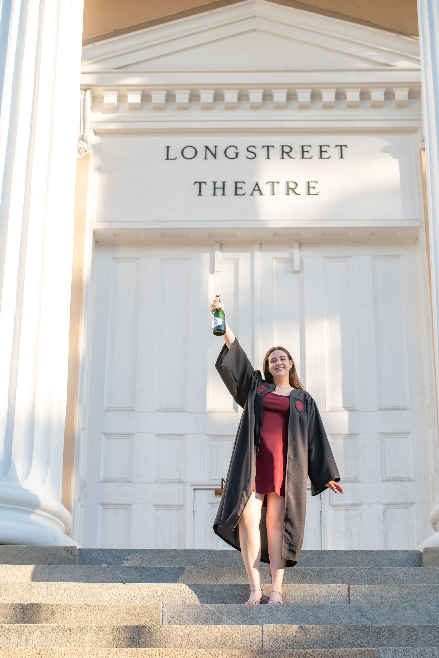 Graduate in cap and gown raises champagne bottle on Longstreet Theatre steps in celebratory pose.