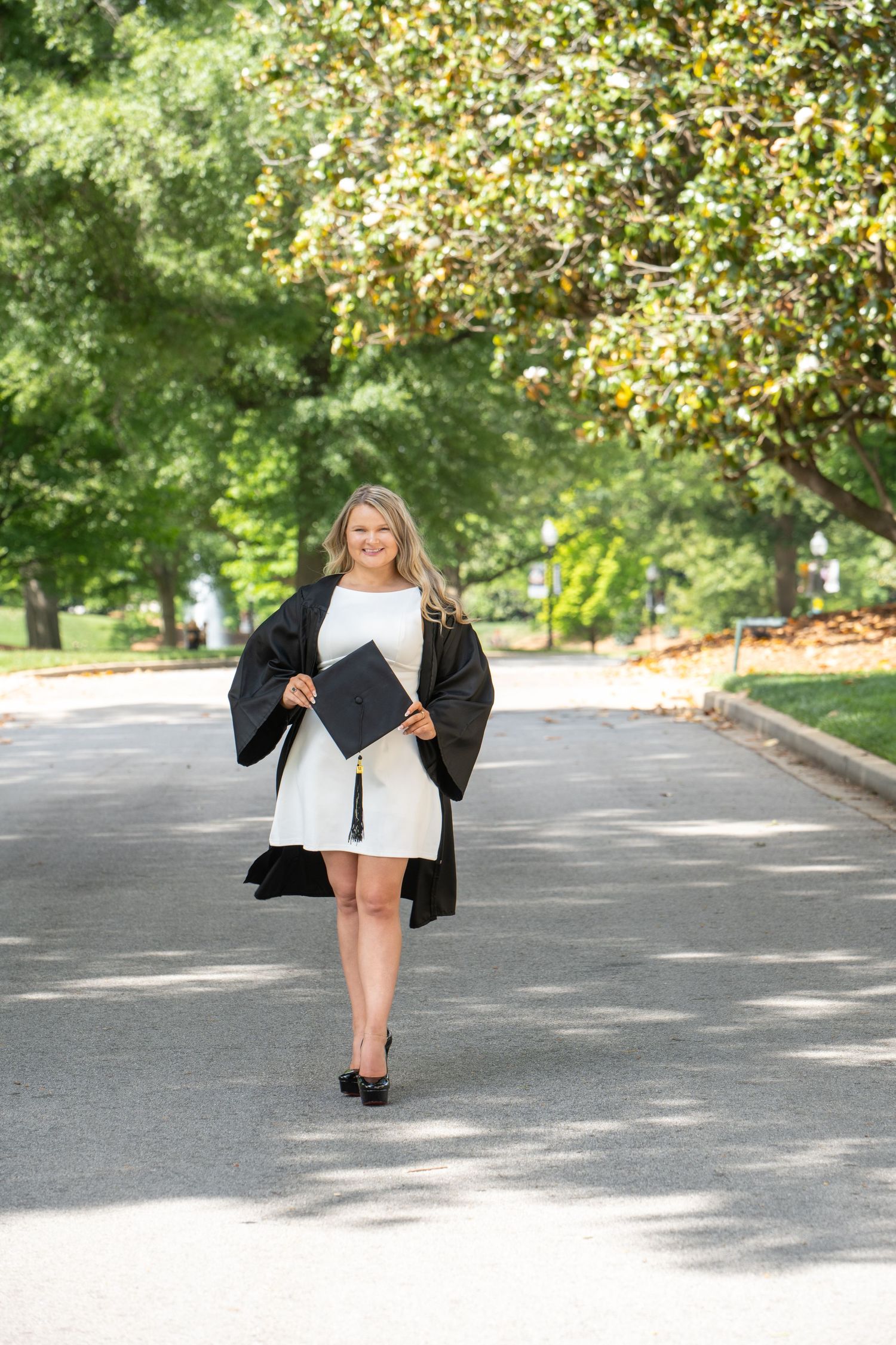 Smiling graduate in black gown holding cap on tree-lined path wearing white dress and heels.