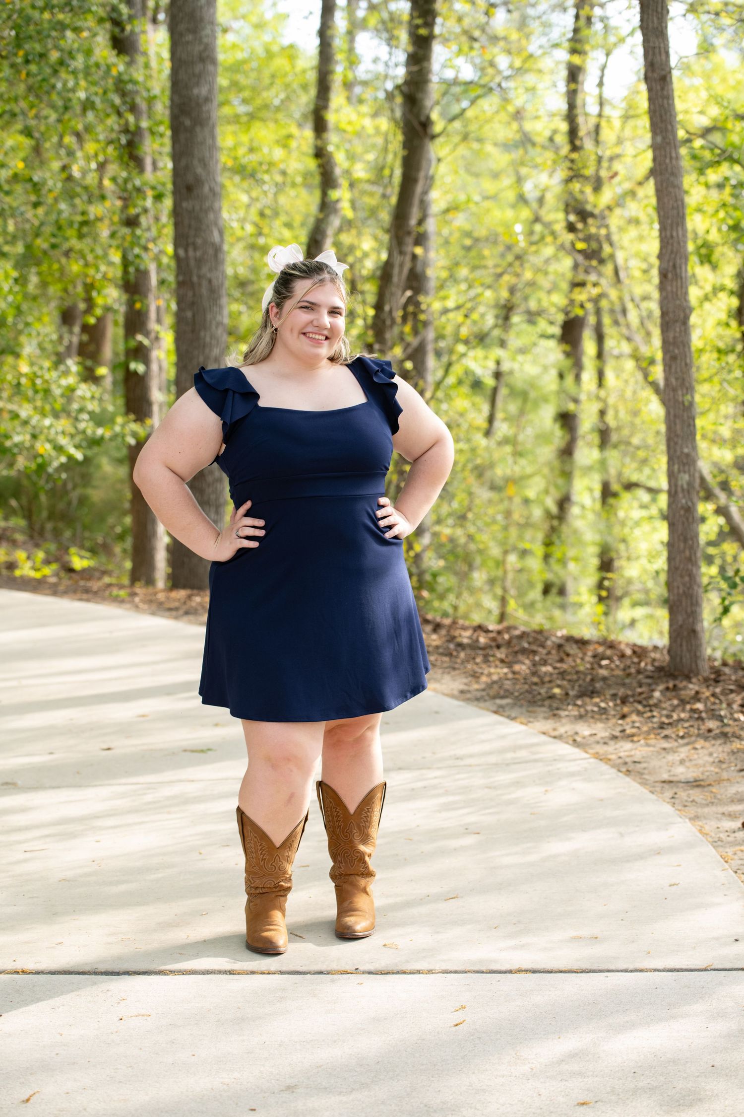Woman in navy dress and cowboy boots posing on a path surrounded by lush green trees in an outdoor setting.