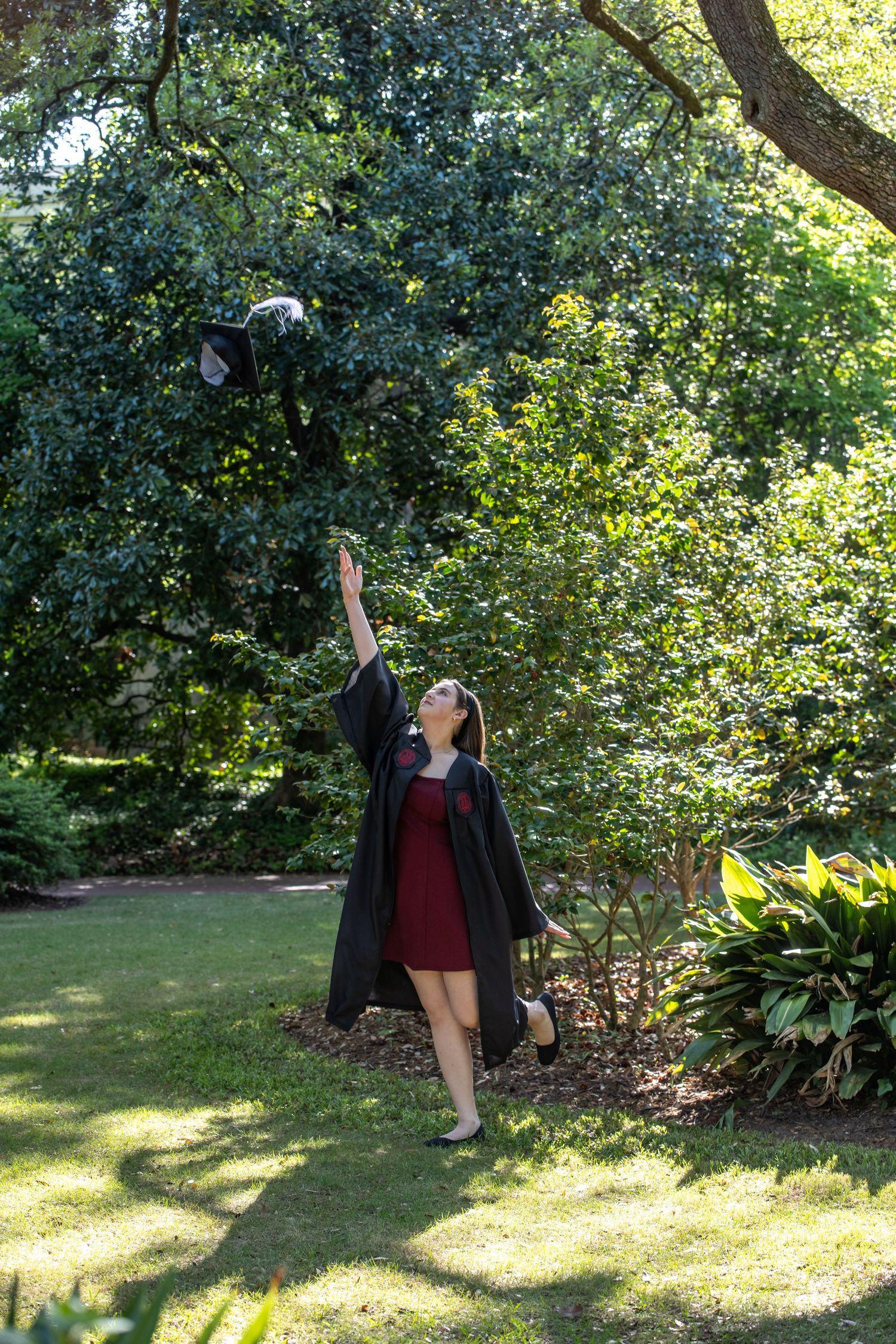 Young woman in graduation gown tossing her cap in the air outdoors in a lush green garden.