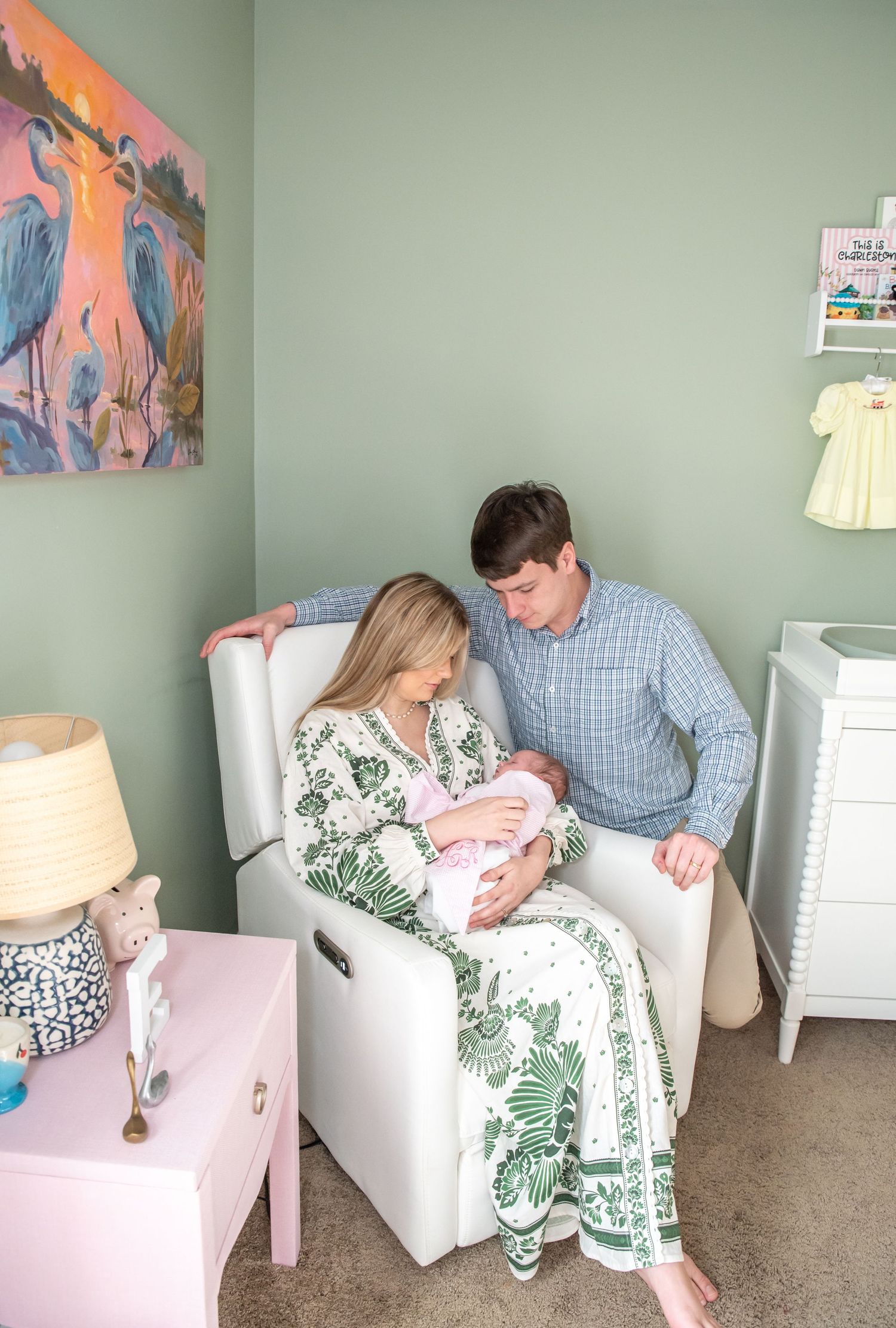 New parents admire their newborn in a white nursery glider, surrounded by sage green walls and soft pink decor.