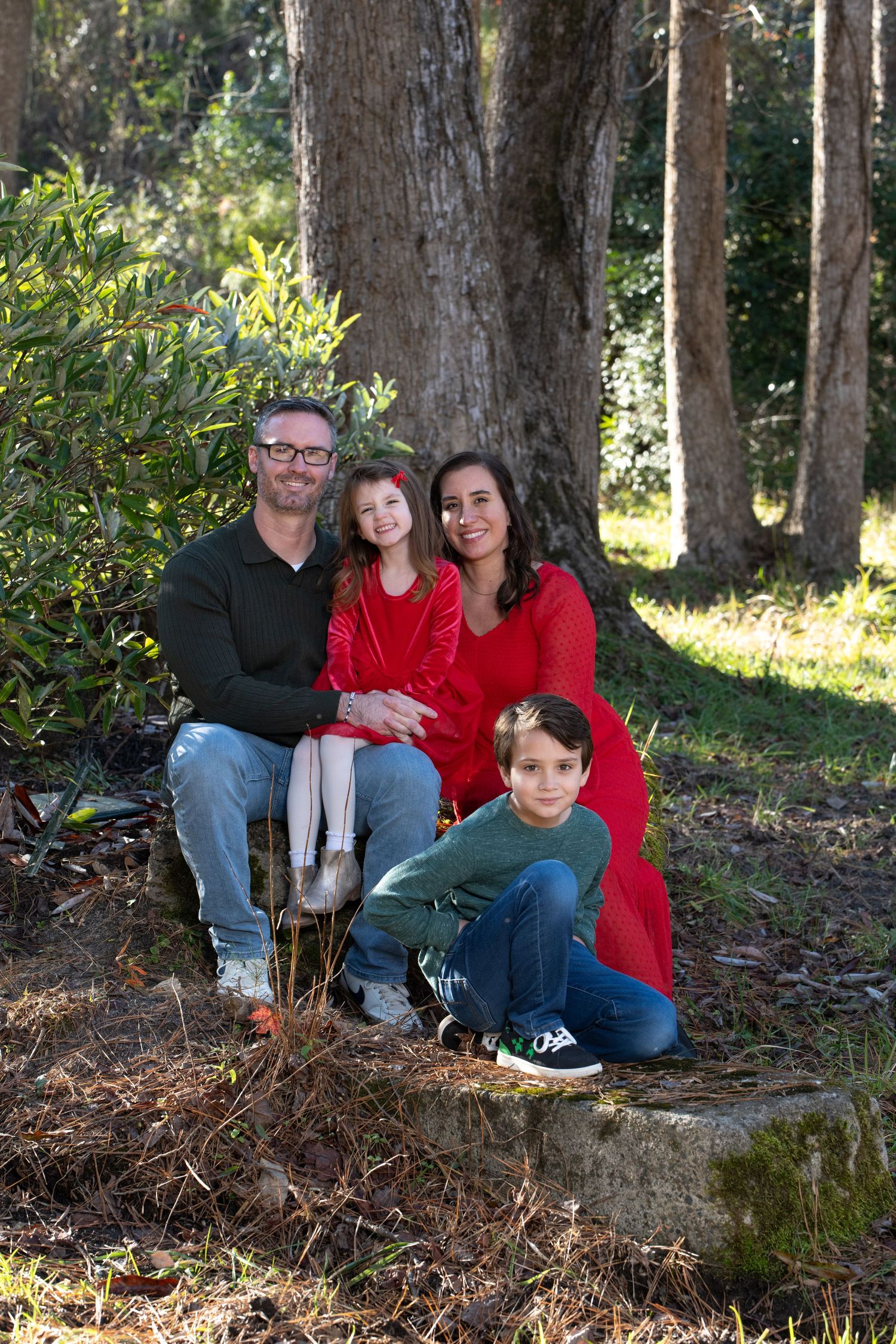 Family of four posing outdoors in a wooded setting, parents and two children wearing red and green for a holiday portrait.