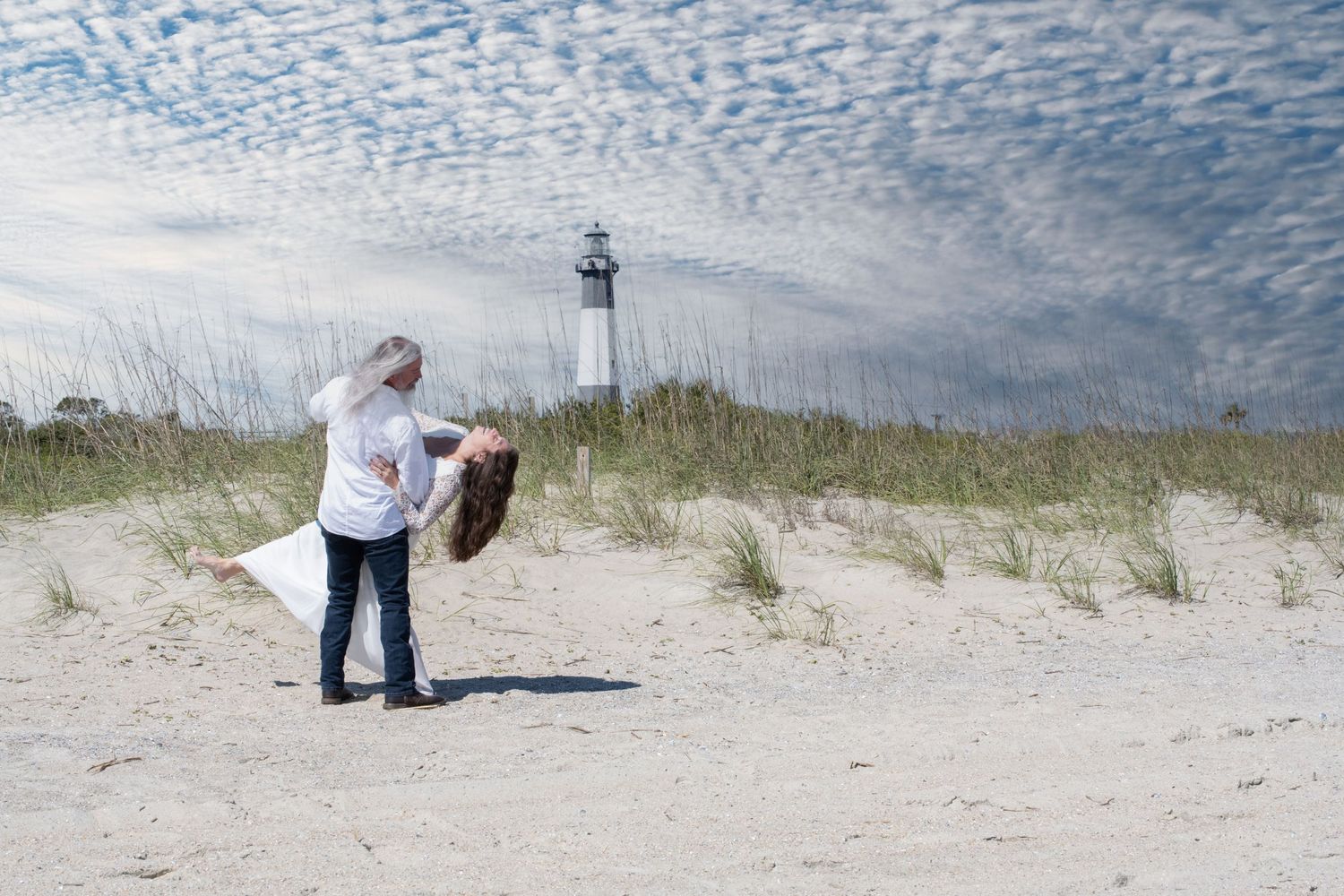 A romantic moment captured on sandy dunes with a lighthouse in the background.