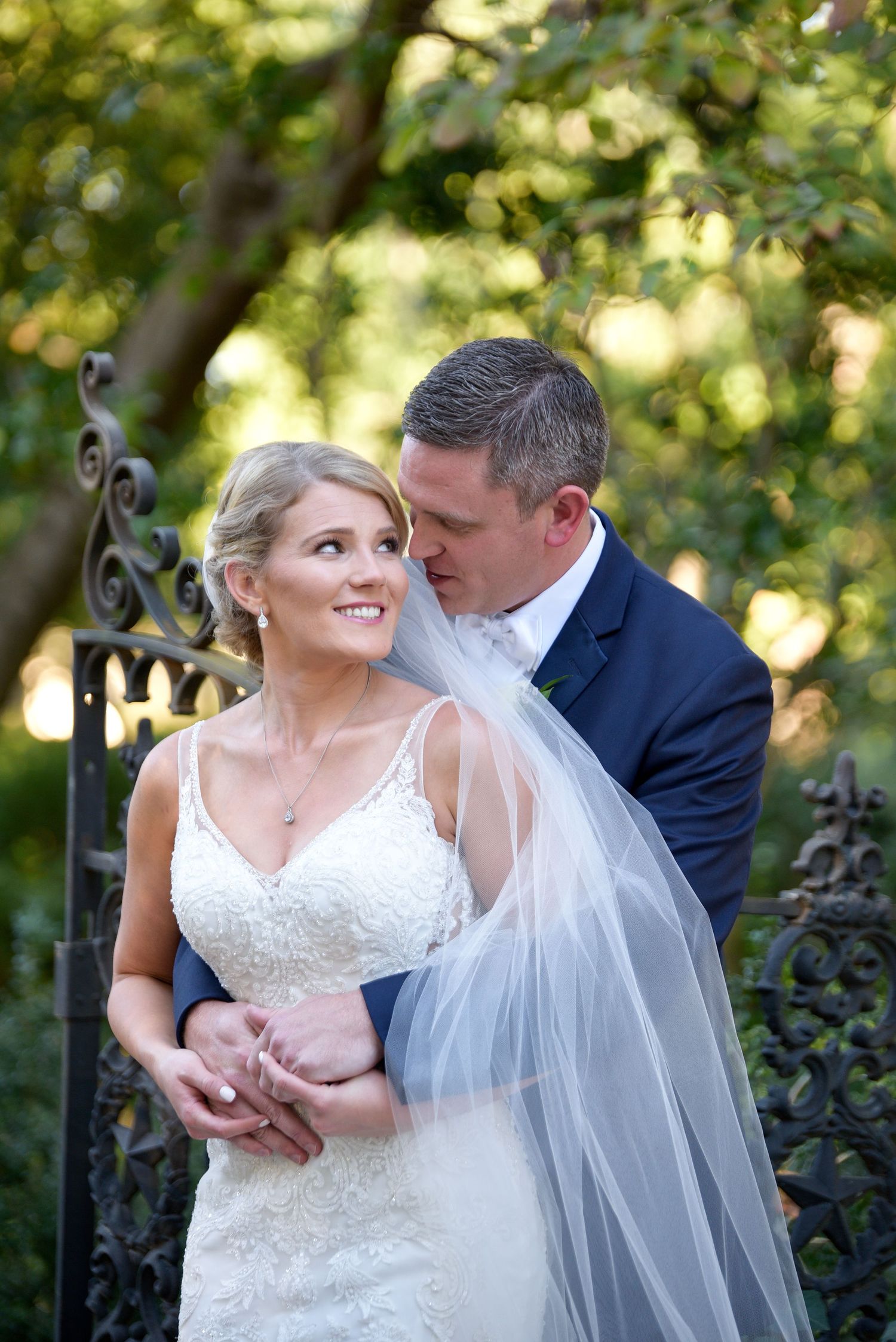 Tender wedding moment captured by ornate garden gate with natural bokeh lighting.