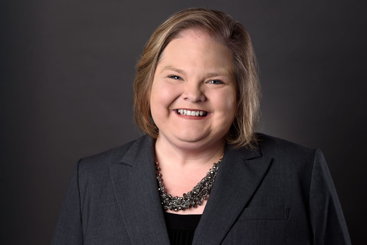 Professional portrait of woman in black blazer smiling warmly against dark studio backdrop.