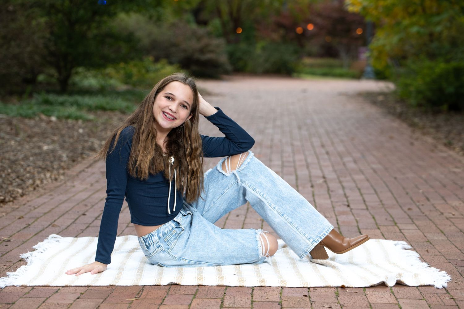 Young woman in navy crop top and ripped jeans sitting on blanket on brick path surrounded by autumn greenery.