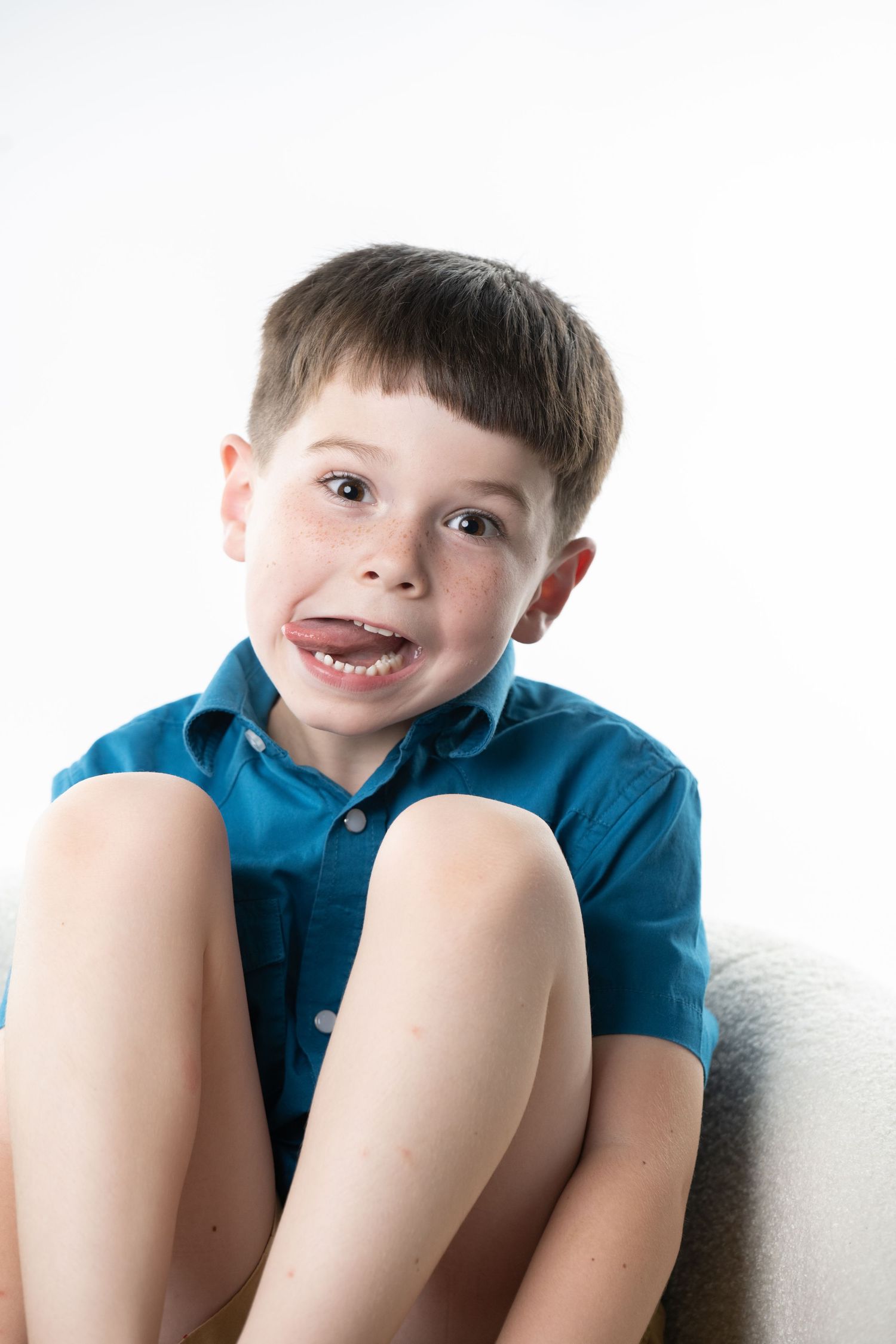 A young boy in a blue shirt sits cross-legged, making a silly face with his tongue out against a white background.