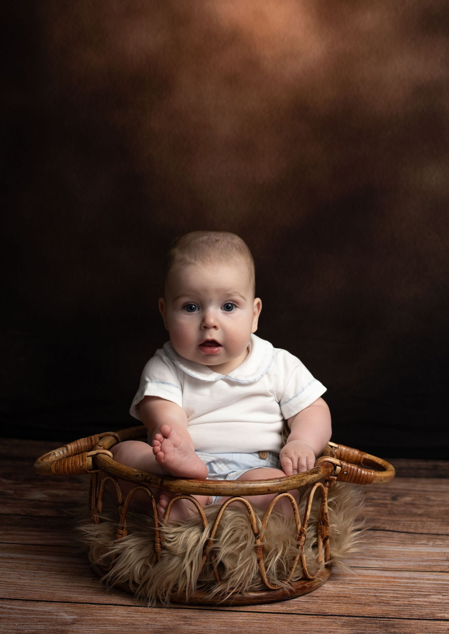 A cute baby sitting upright in a wicker basket with fur lining on a wooden floor against a brown backdrop.