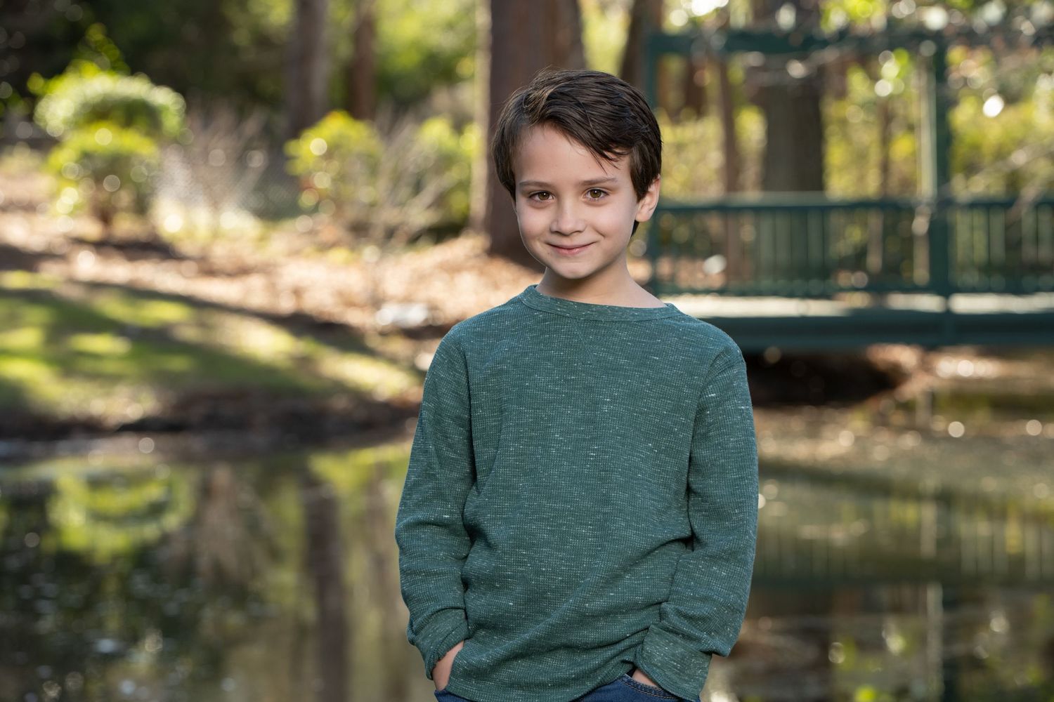 A smiling boy in a green sweater stands with hands in pockets near a pond in a lush park setting.