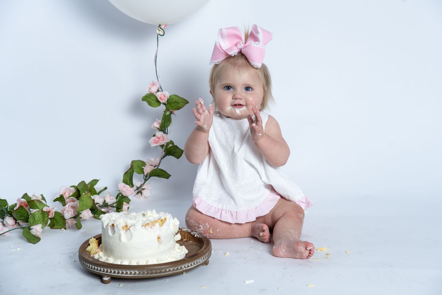 Baby in white top and pink bow sits next to first birthday smash cake and climbing vine against white backdrop.
