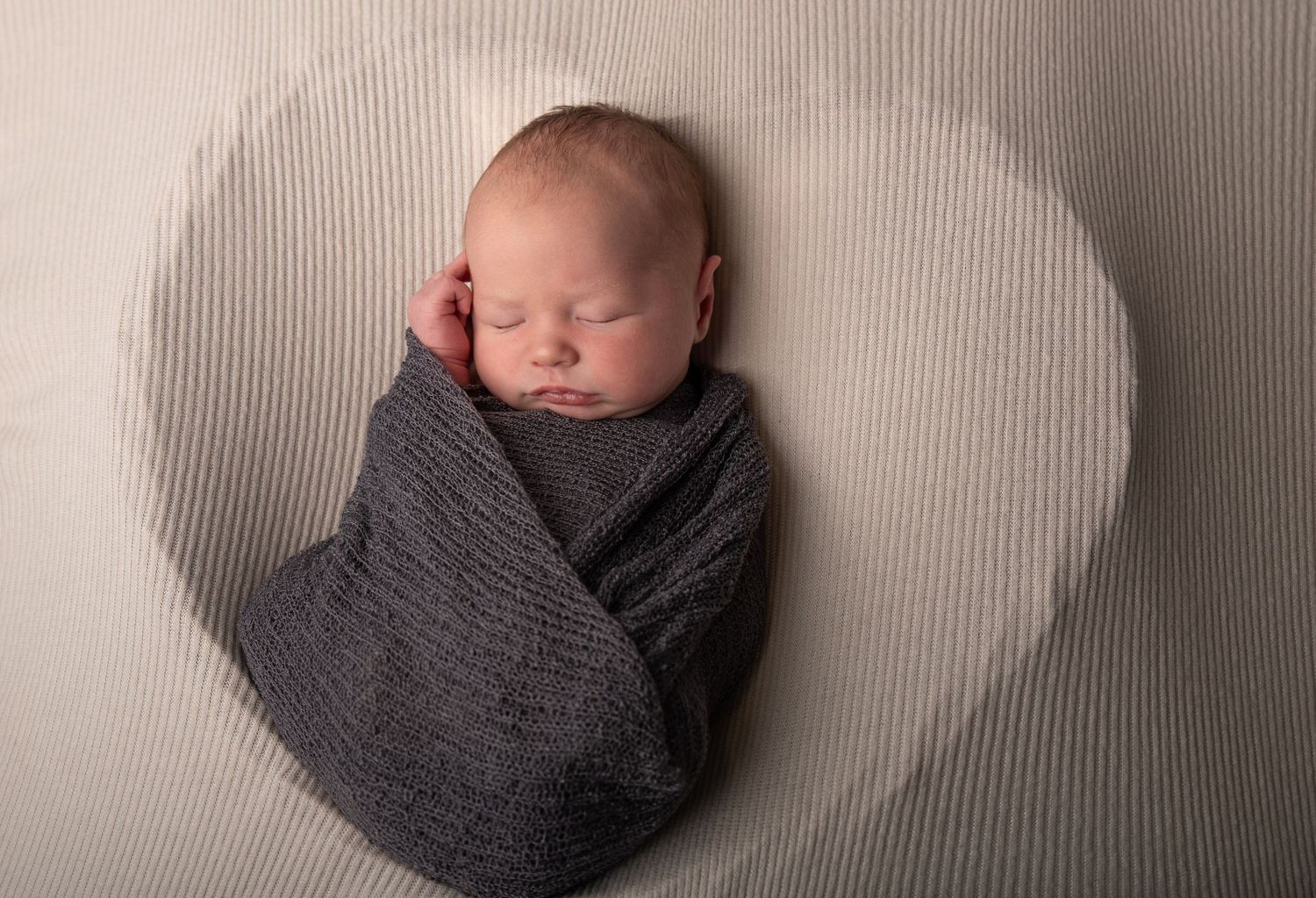 Newborn baby swaddled in gray wrap sleeping peacefully on a cream background with a subtle heart design.