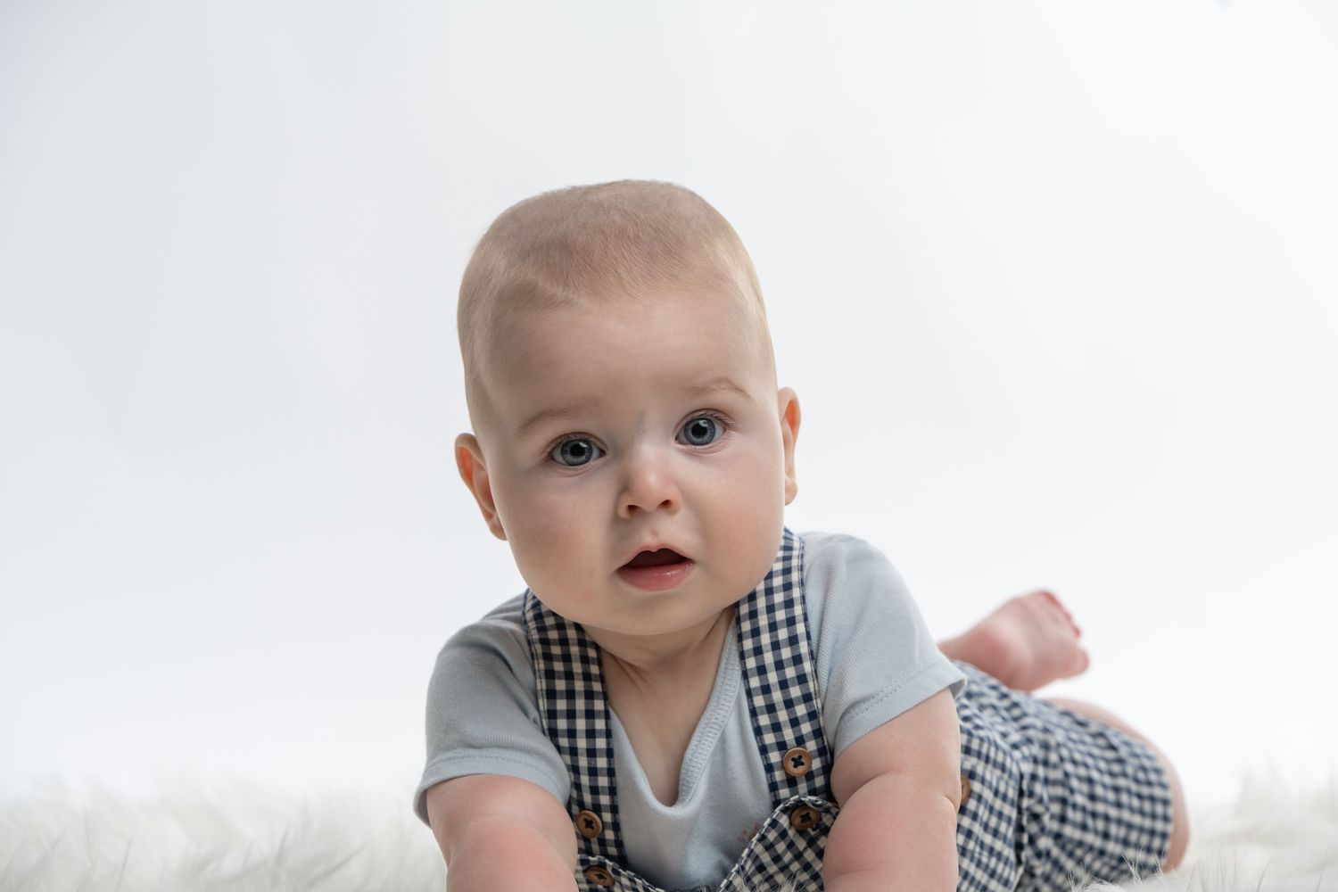 Adorable baby in gingham overalls doing tummy time on a white fluffy rug against a light background.