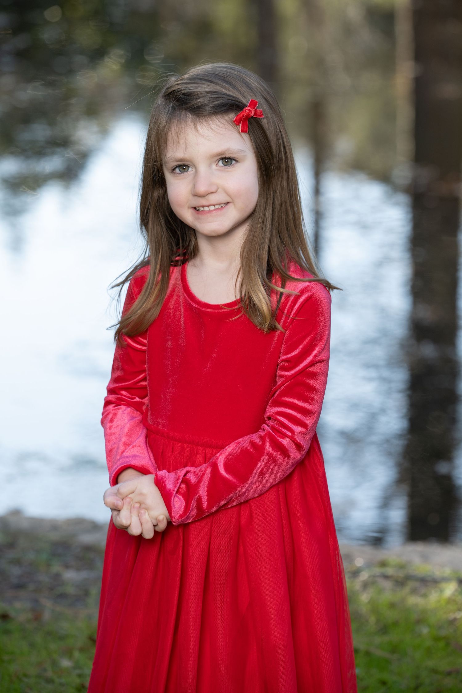 Young girl in red velvet dress with matching bow smiles outdoors near a reflective pond.