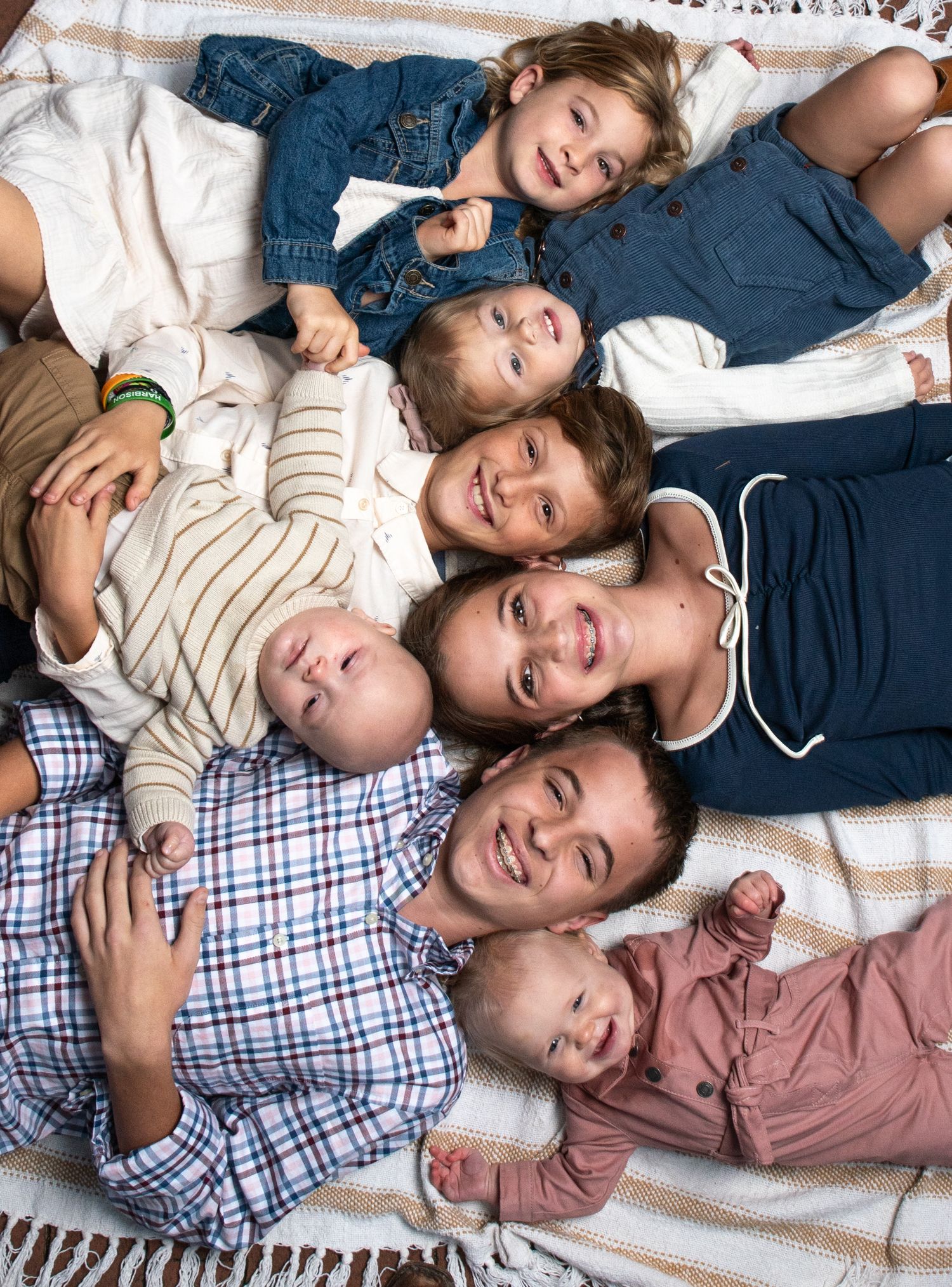 Family of seven lying together on a blanket, including parents, baby, toddlers, and young children, smiling warmly.